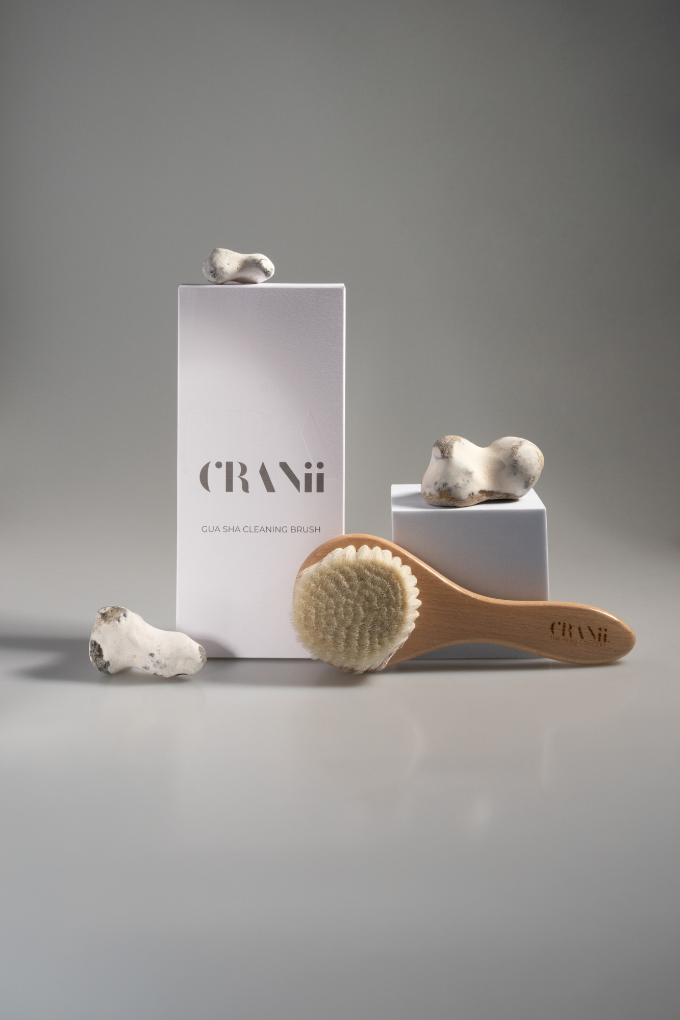 Still-life product photo of a CRANii gua sha cleansing brush with white packaging and sculptural stones on a gray studio background.