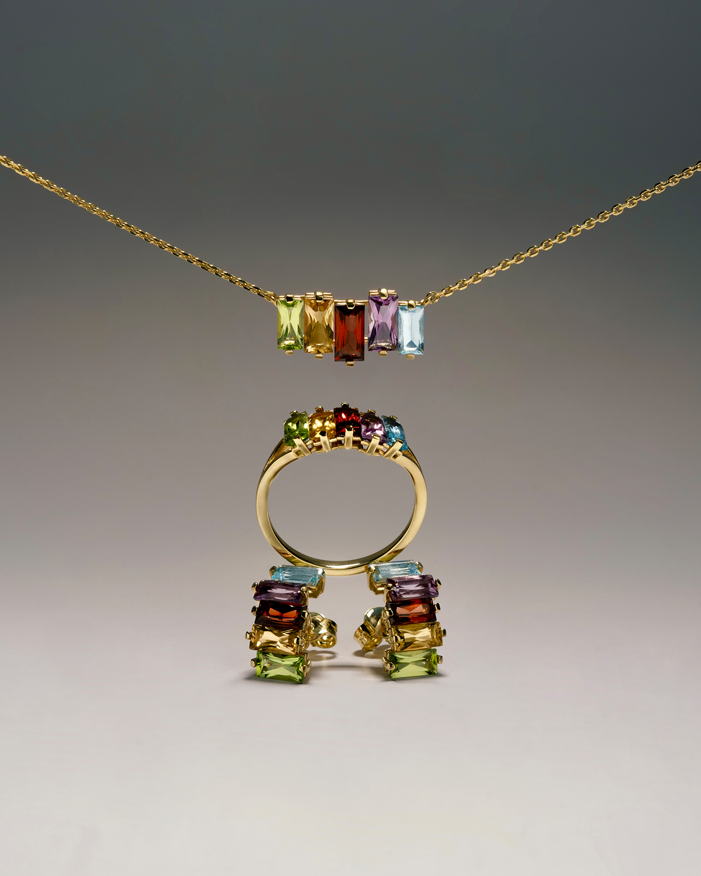 Gold jewelry set with colorful rectangular gemstones, including a necklace, ring, and earrings, arranged on a neutral studio background.