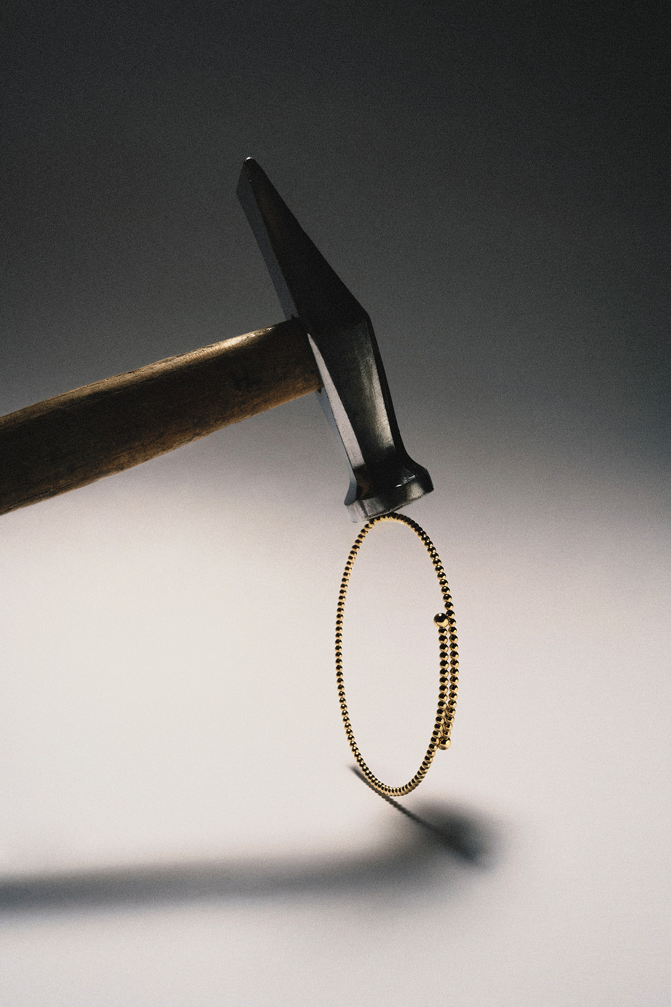 Gold bracelet suspended beneath a hammer in a dramatic studio still life.