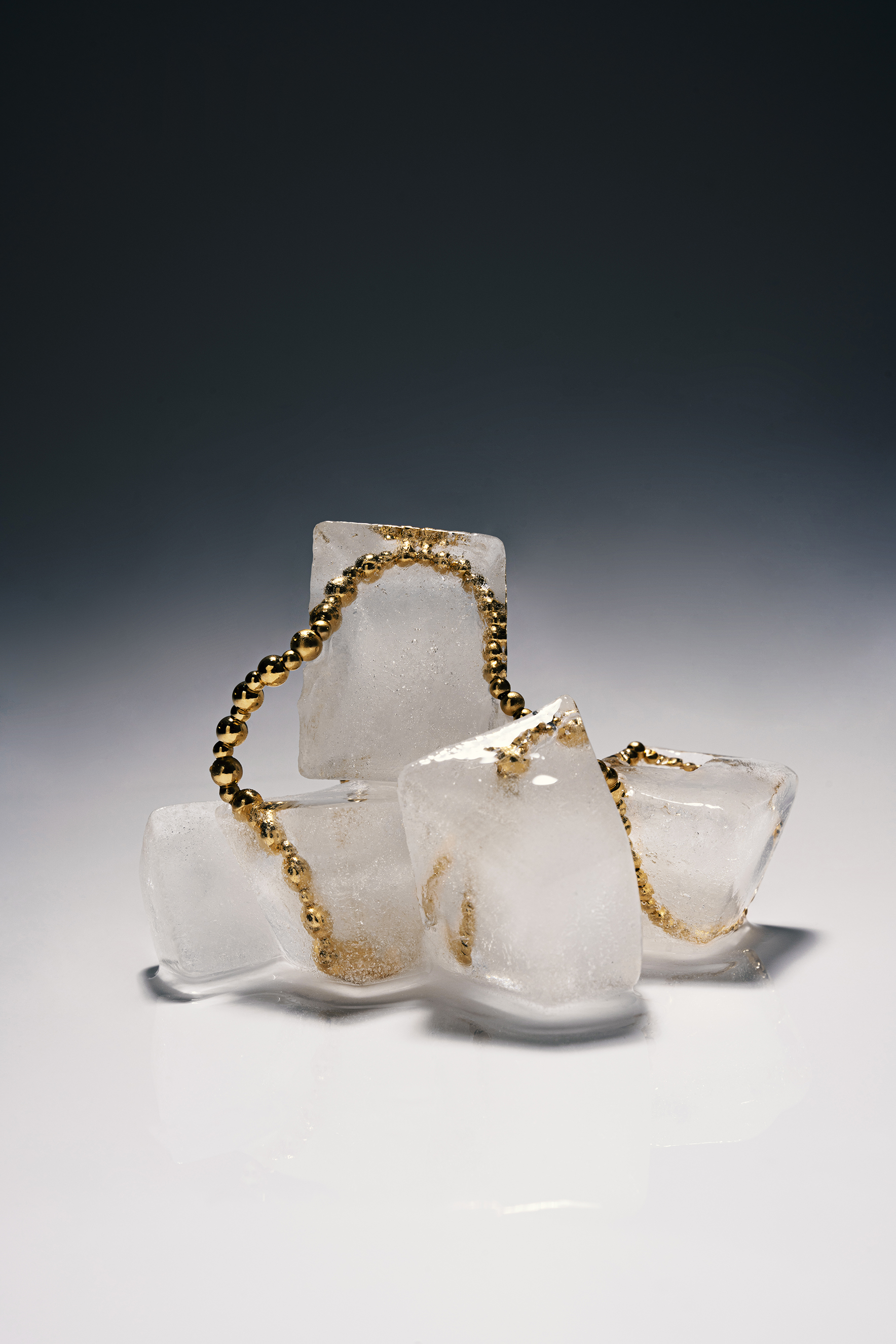 Gold beaded bracelet arranged around melting ice cubes on a reflective studio surface.