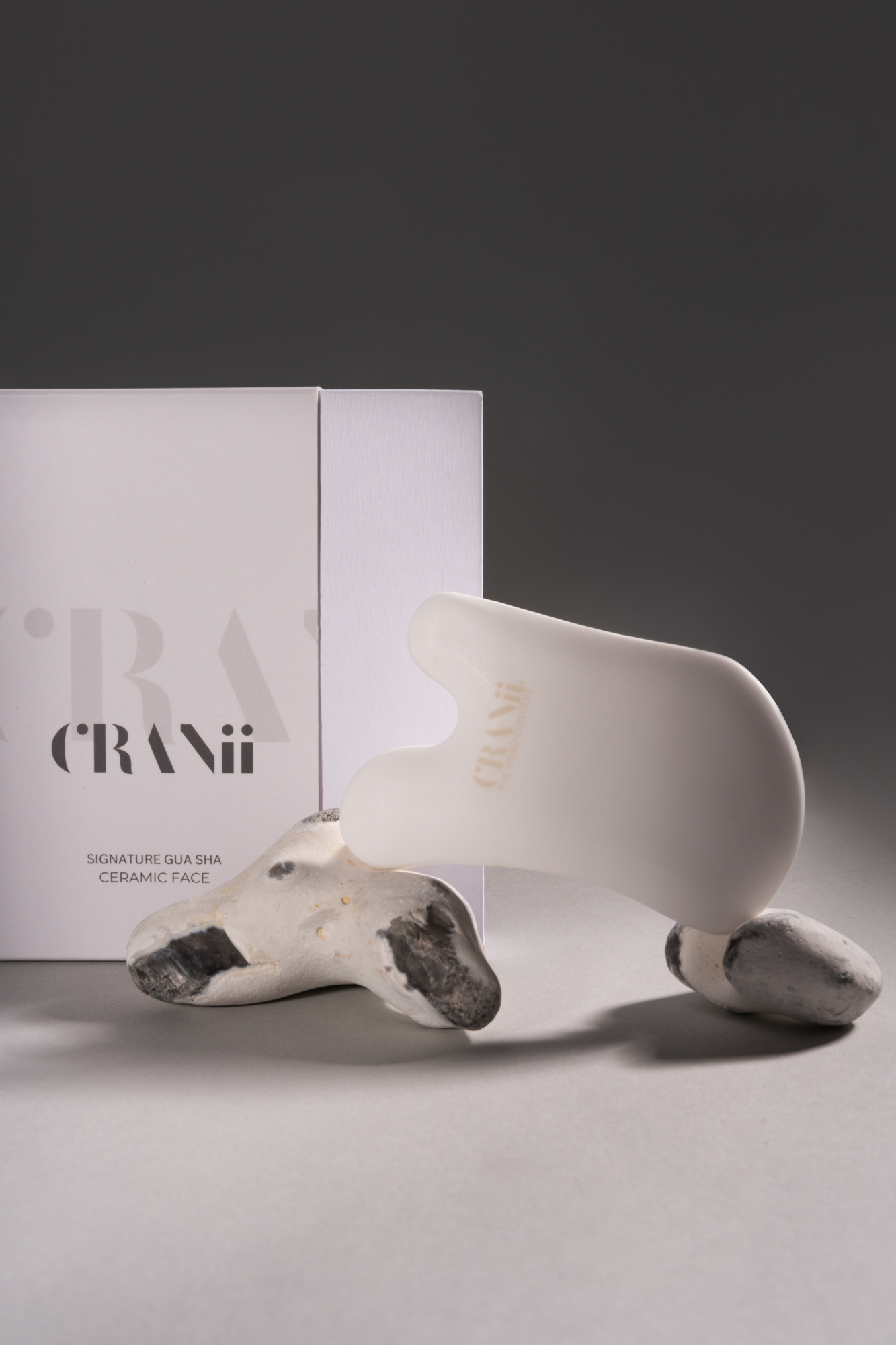 White CRANii ceramic face tool displayed with product packaging and sculptural stones on a gray studio background.