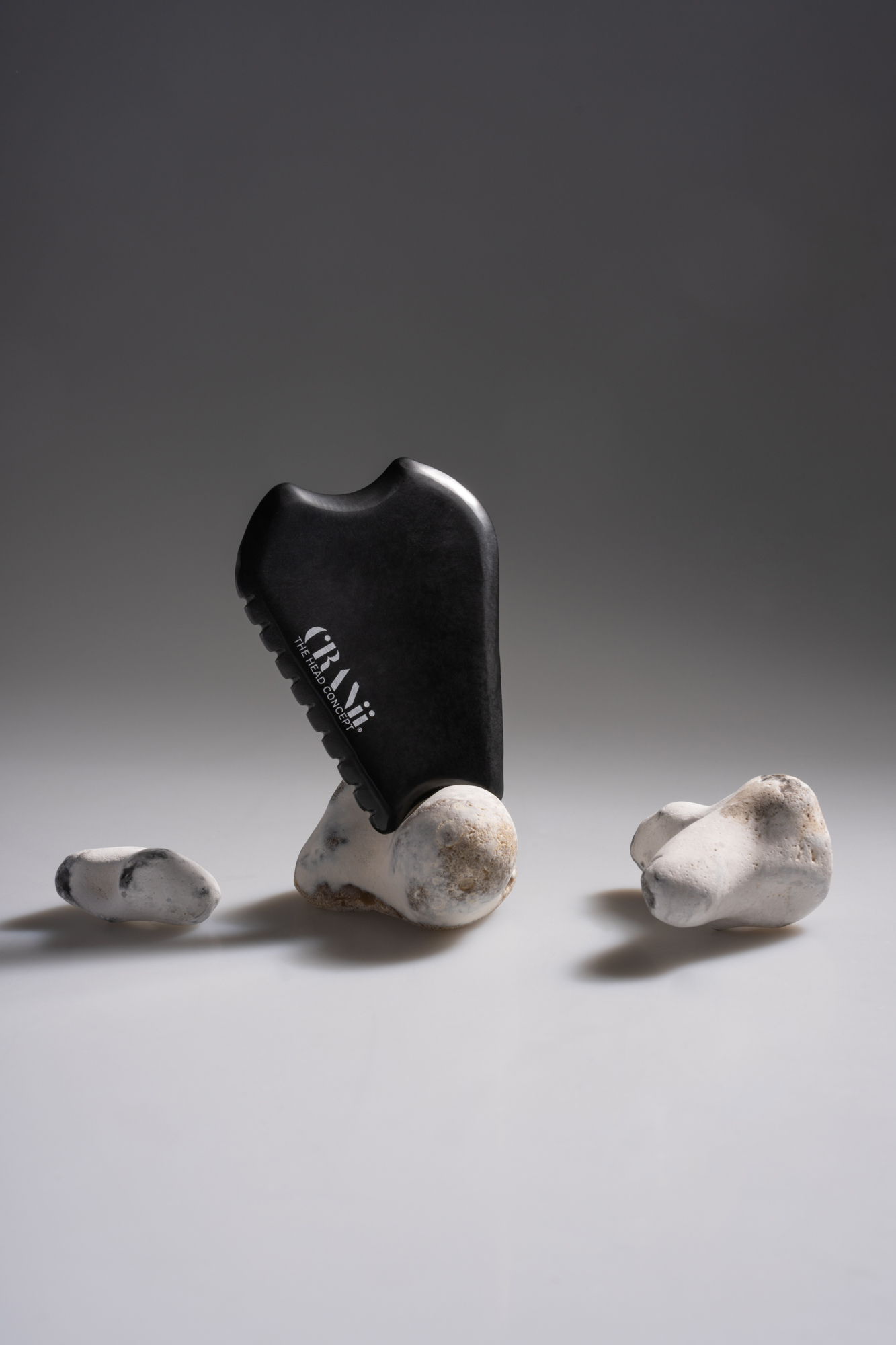Black CRANii ceramic face tool balanced on white stones in a minimalist studio setup.