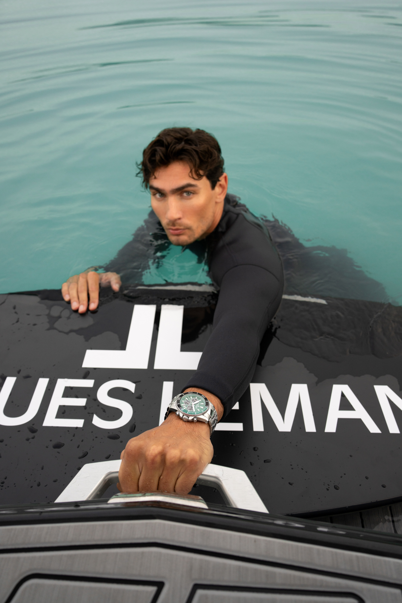Man in a black wetsuit holding onto a boat from the water, wearing a silver watch with a green dial.