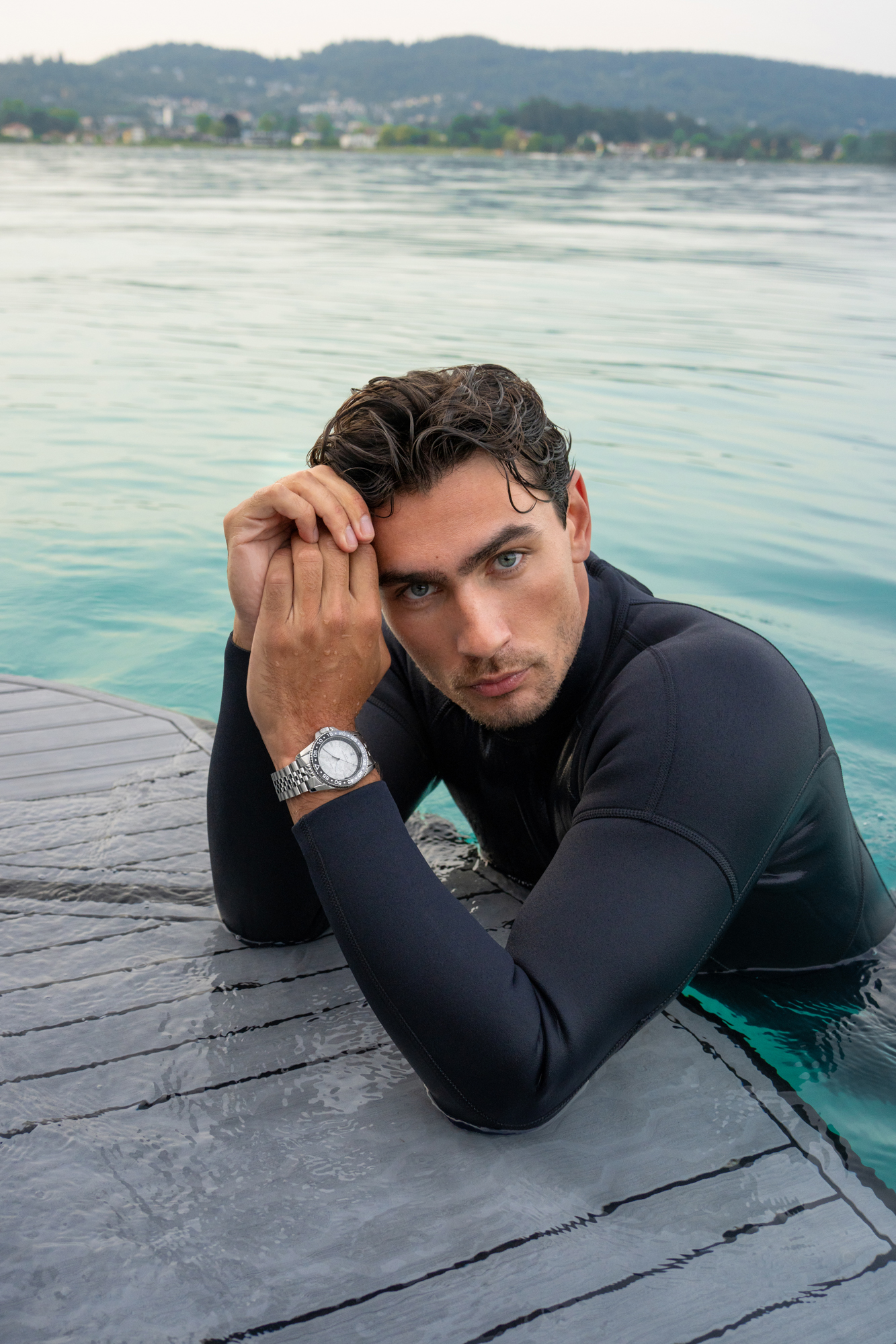 Man in a black wetsuit leaning on a dock in turquoise water, wearing a silver wristwatch.