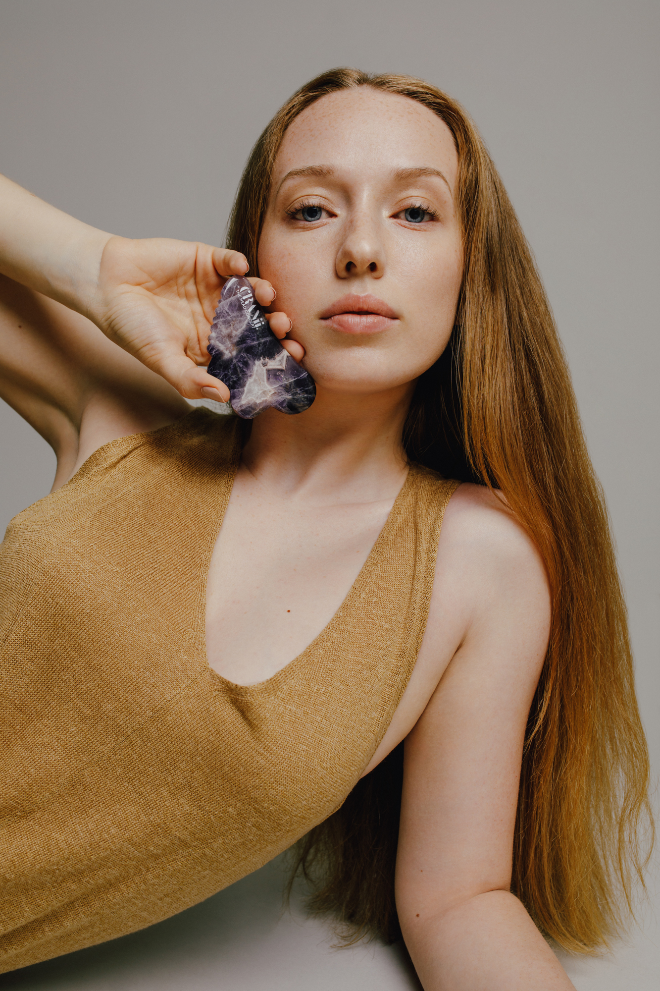 Red-haired model in a gold top holding a purple crystal against her cheek in a studio portrait.