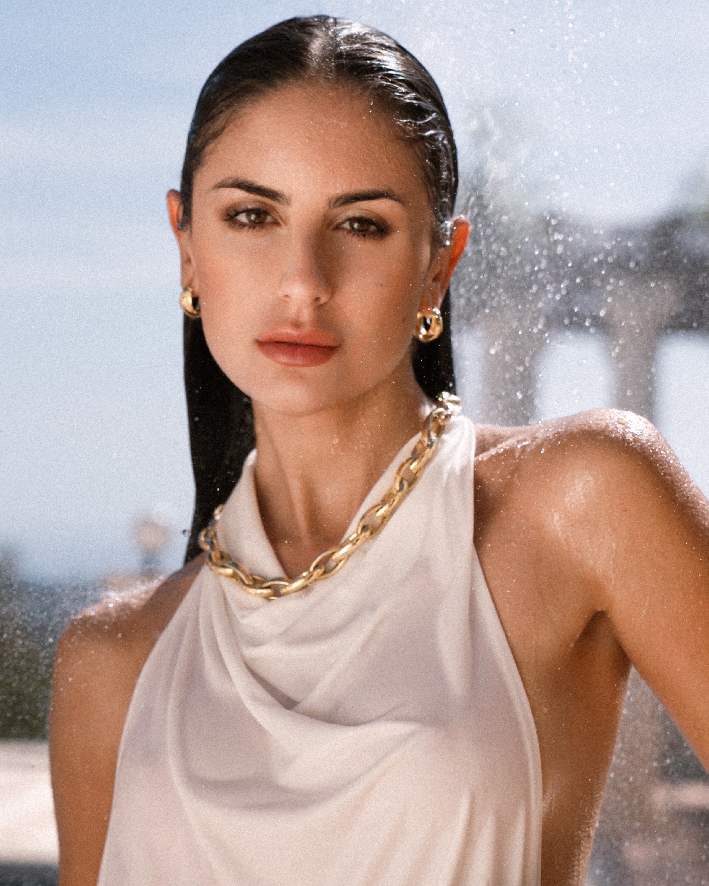 Close-up portrait of a woman in a white halter top wearing gold jewelry, photographed with water spray in bright sunlight.