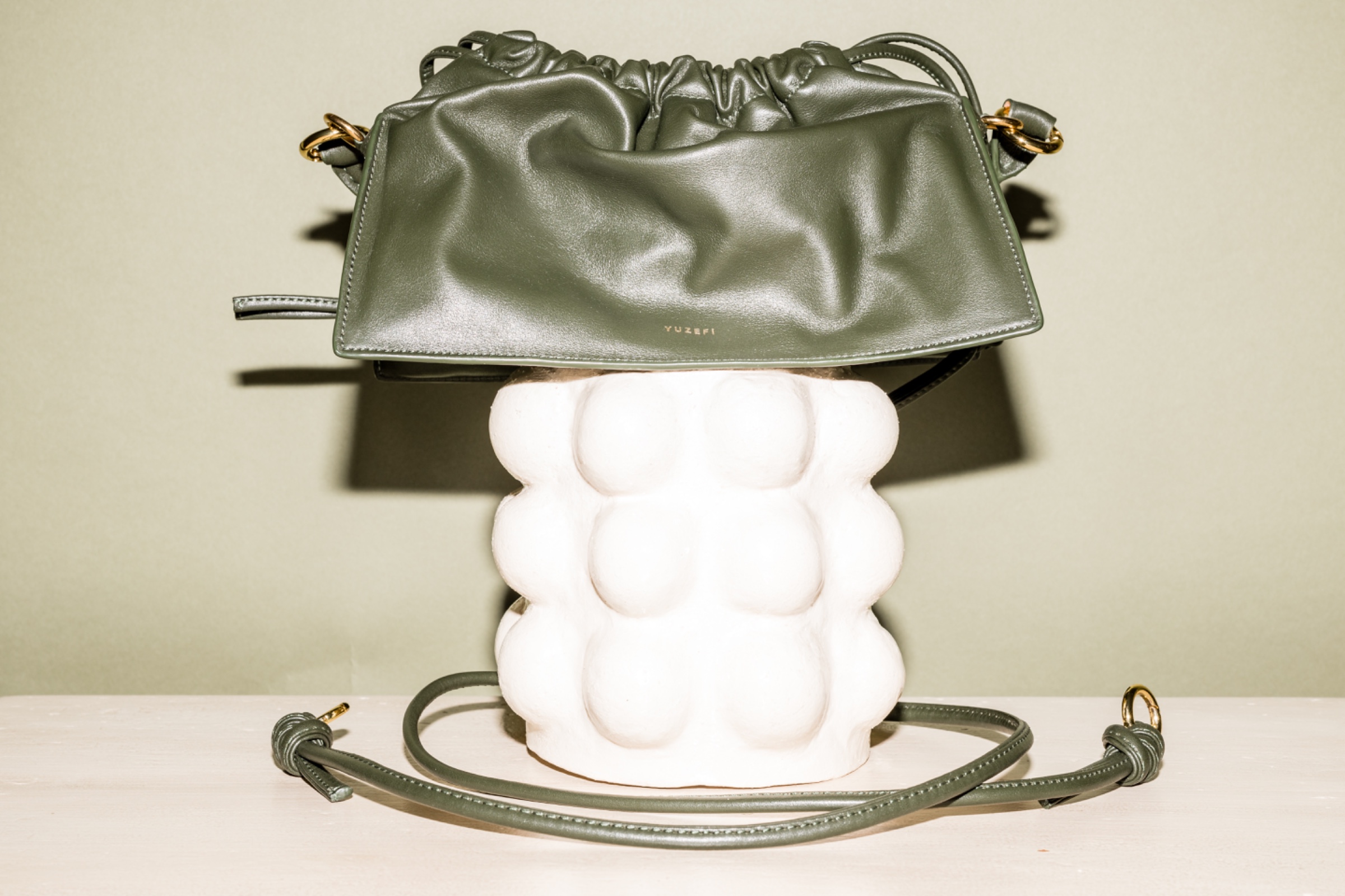 Olive green leather handbag displayed on a white sculptural pedestal in a studio product shot.