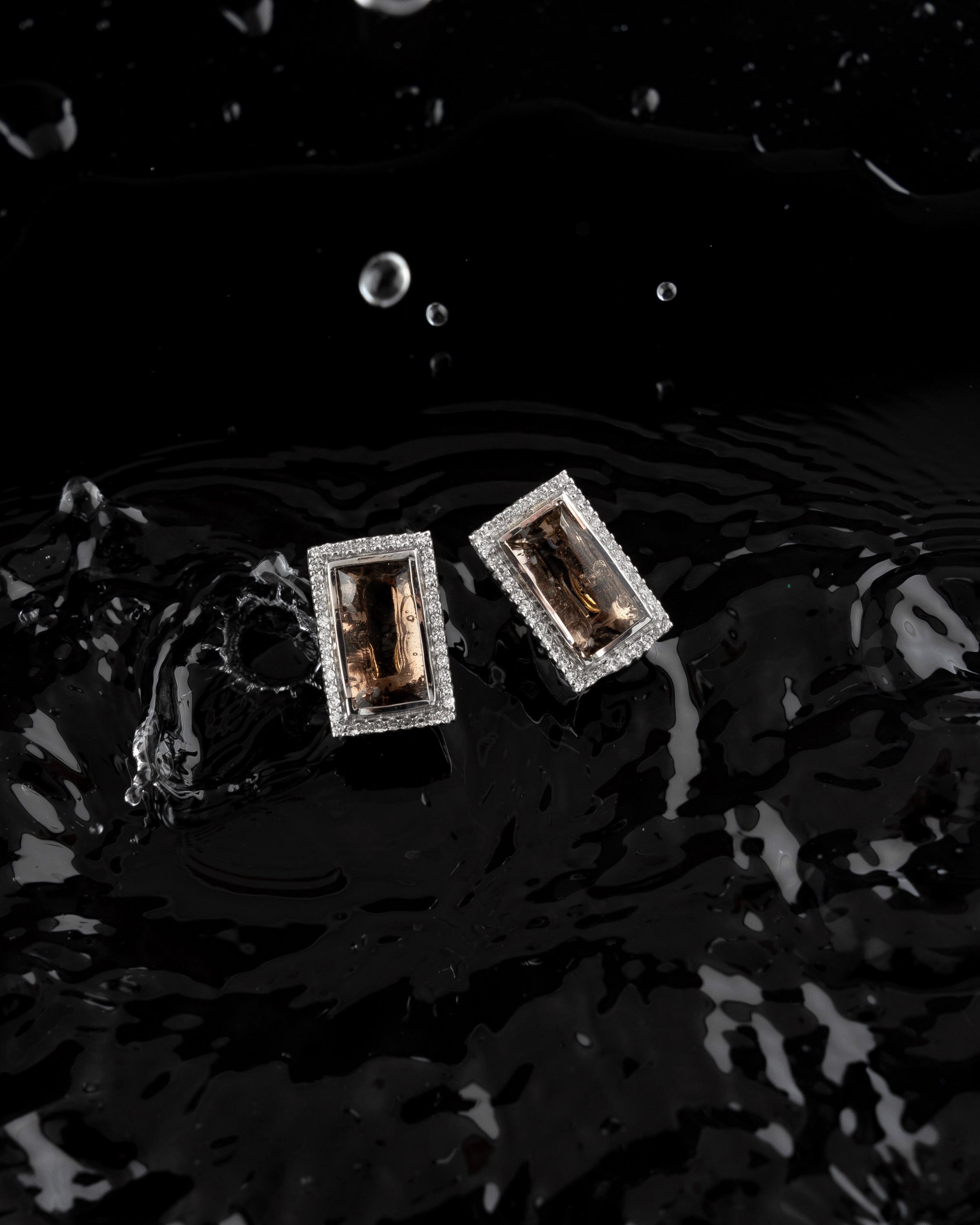 Pair of rectangular gemstone earrings with sparkling borders placed on a glossy black liquid surface.
