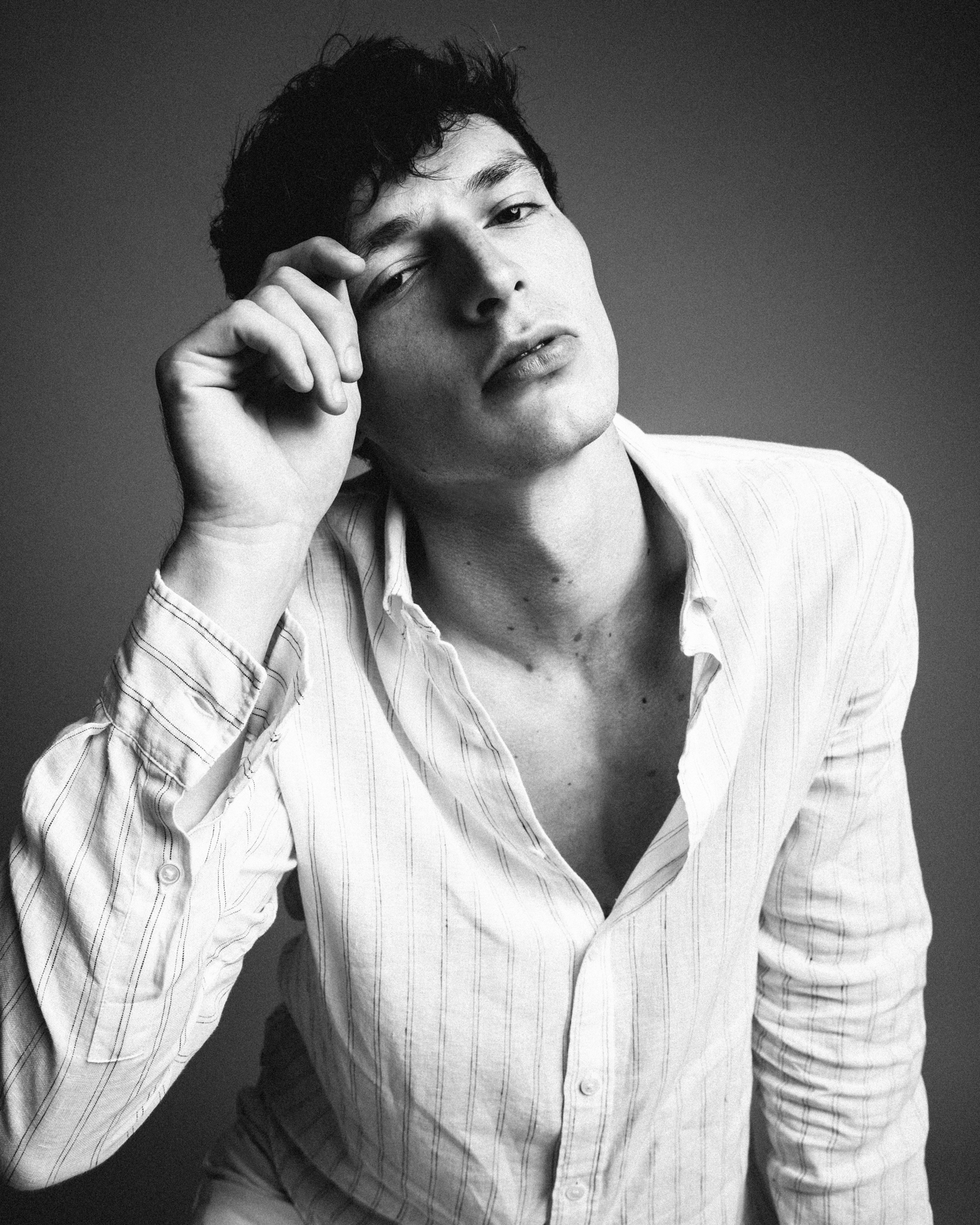 Black-and-white studio portrait of a man in an open white shirt resting one hand near his face.