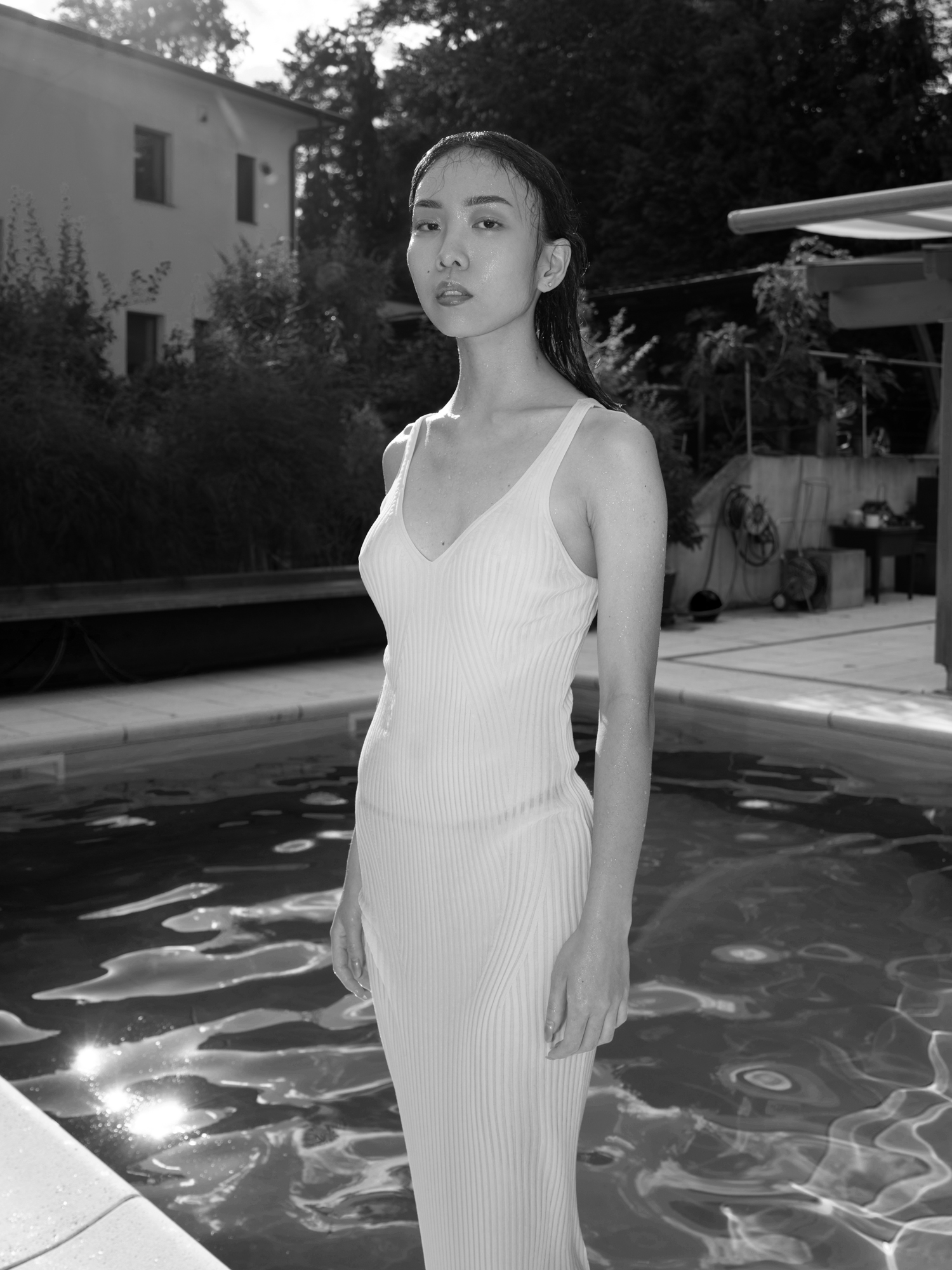 Black-and-white portrait of a woman in a white ribbed dress standing beside a swimming pool.