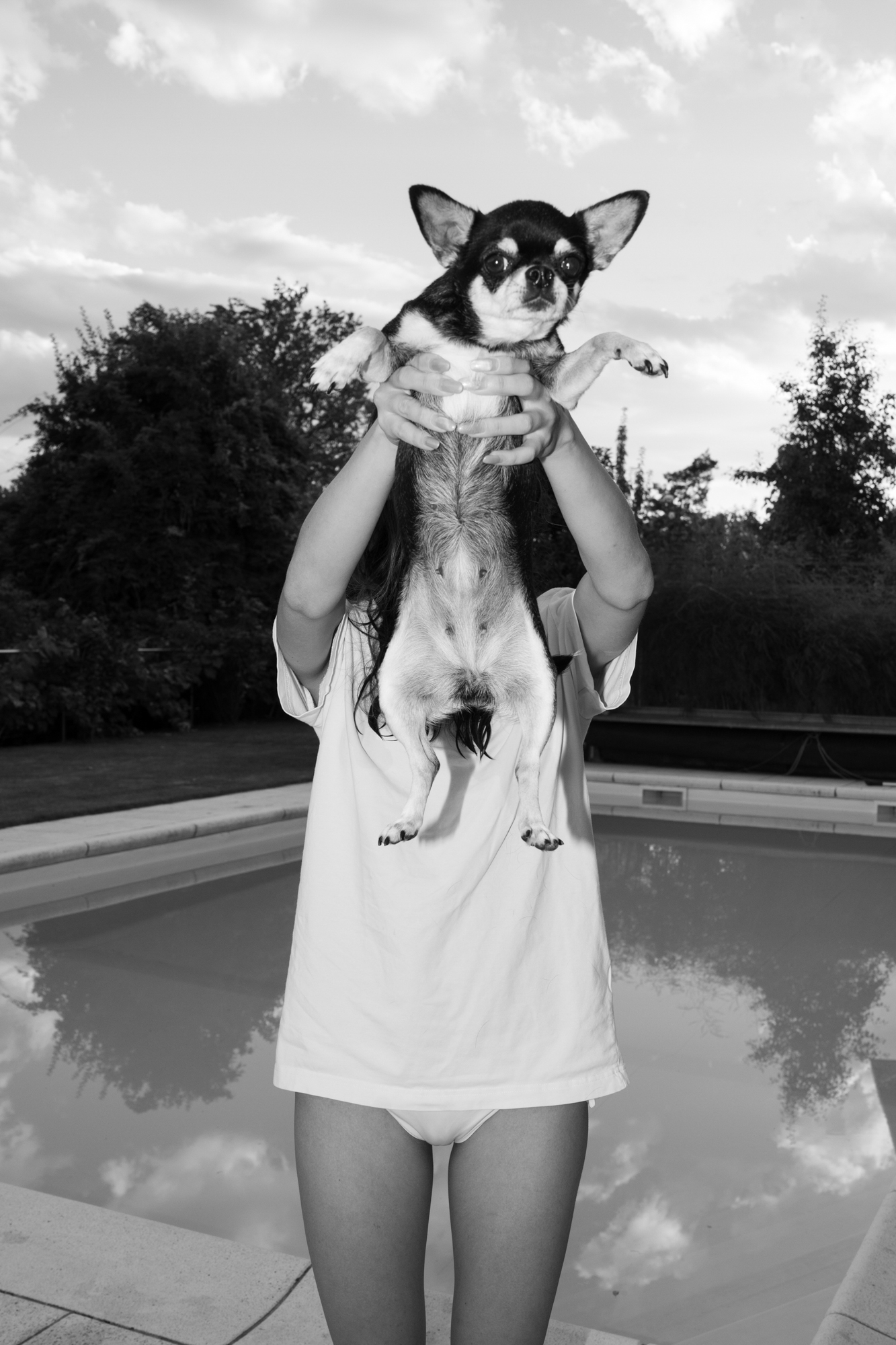 Black-and-white photo of a person holding a small dog up in front of their face by a swimming pool.