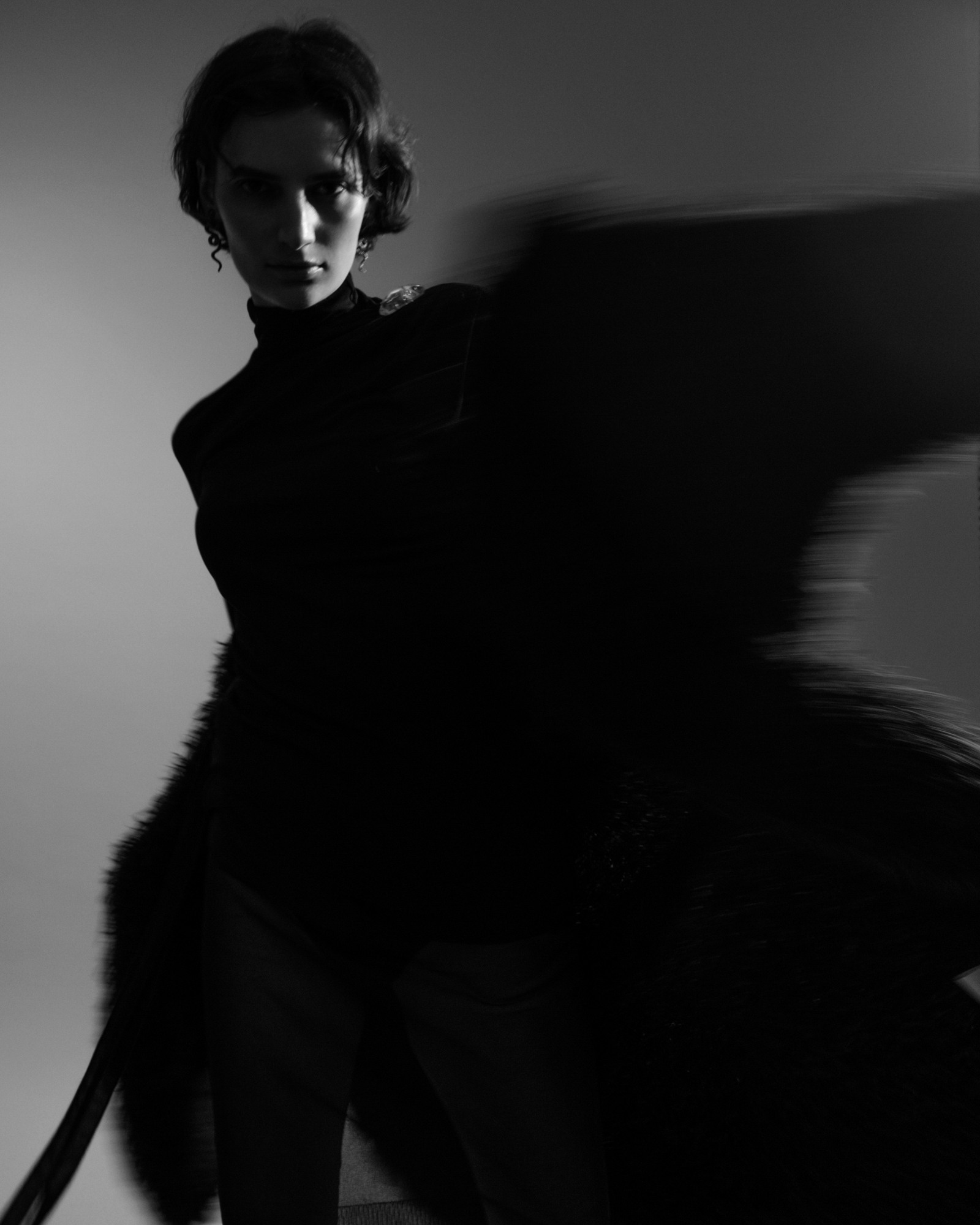 Black-and-white fashion portrait of a model in dark clothing with a blurred fur-like garment in motion.