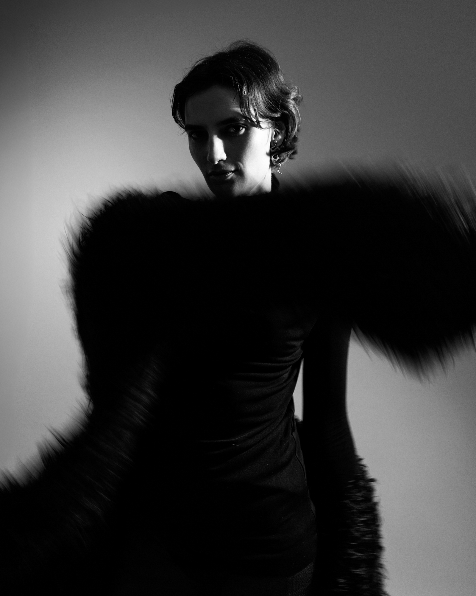 Black-and-white fashion portrait of a model in dark clothing with a dramatic fur-like accessory moving across the frame.