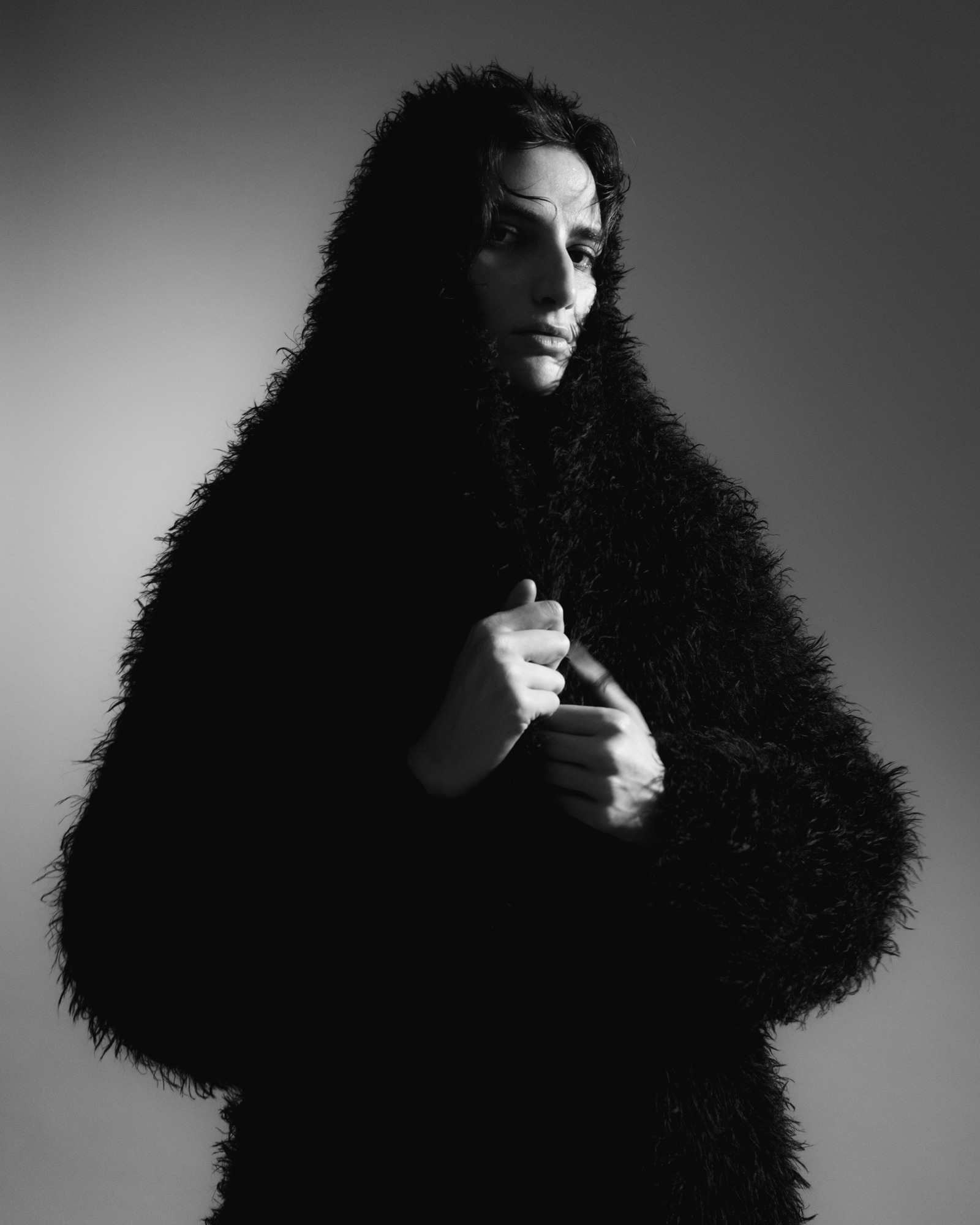 Black-and-white portrait of a model wrapped in a voluminous dark textured coat against a studio background.