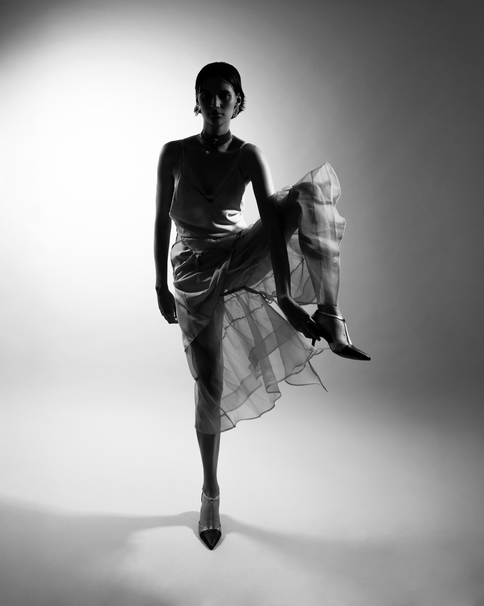 Black-and-white full-body fashion portrait of a model posing in a sheer layered outfit and pointed heels.