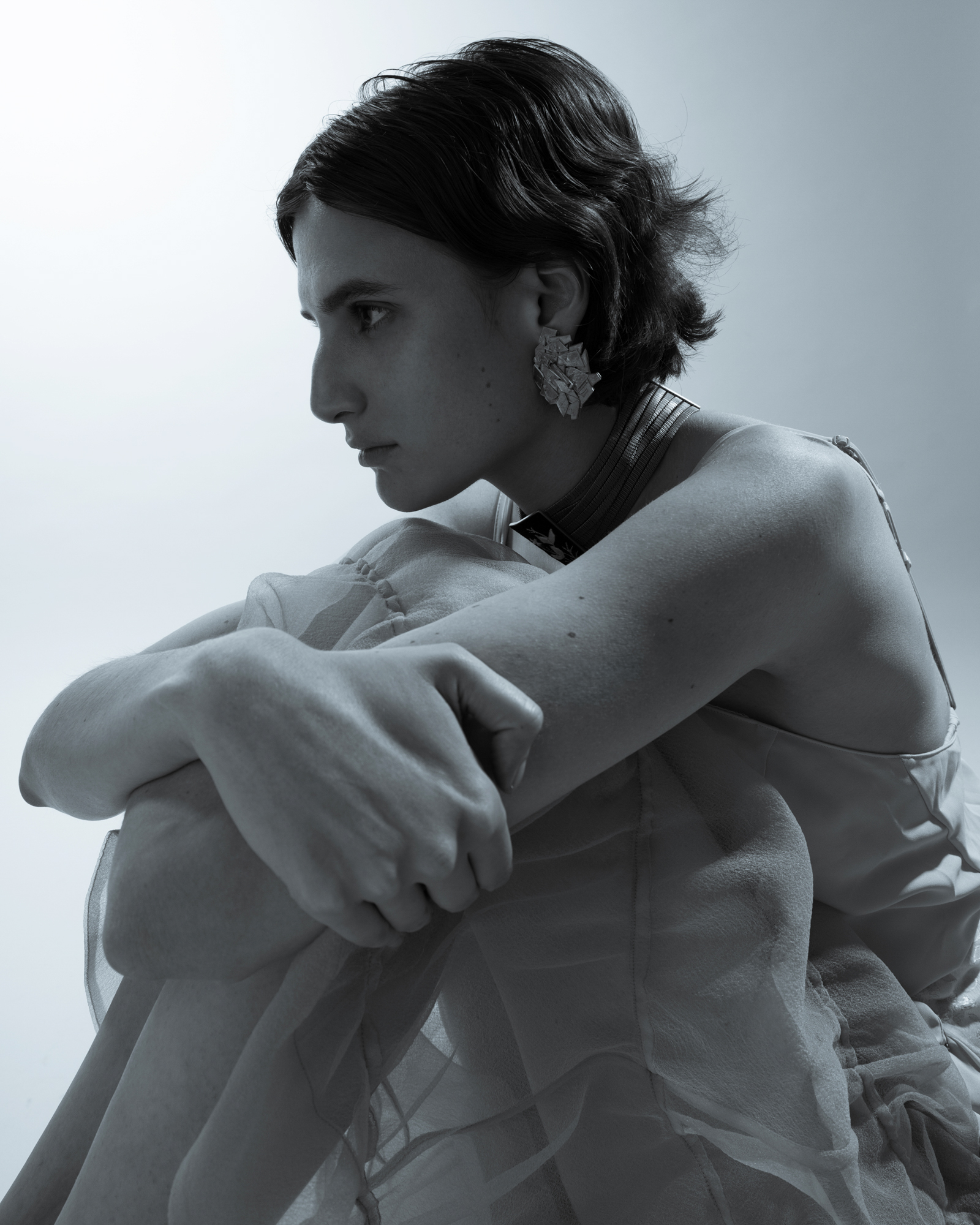 Black-and-white fashion portrait of a seated woman in profile, wearing a sheer textured top and statement earrings.