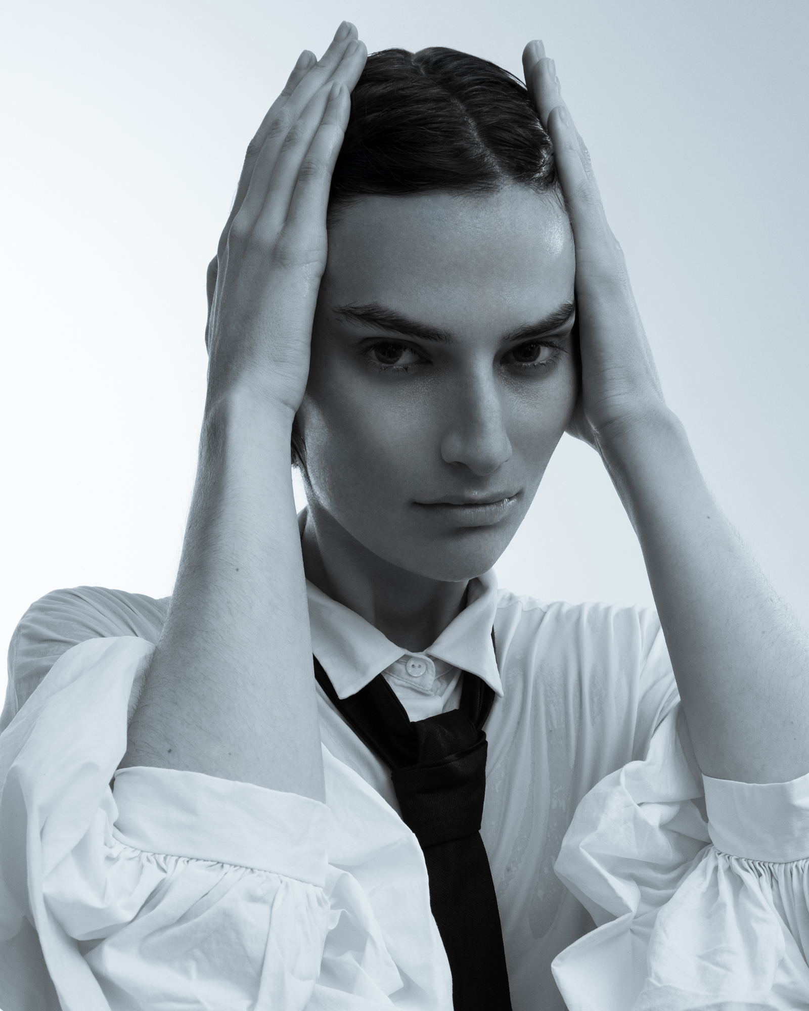 Black-and-white fashion portrait of a woman in a white shirt and black tie, holding her hands against the sides of her head while looking at the camera.