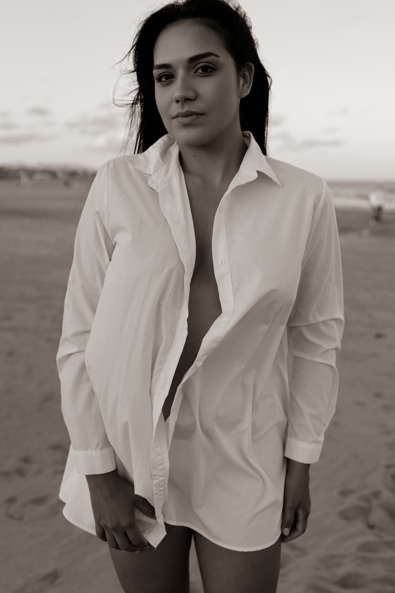 Black-and-white portrait of a woman on a beach wearing an oversized white button-down shirt and looking at the camera.