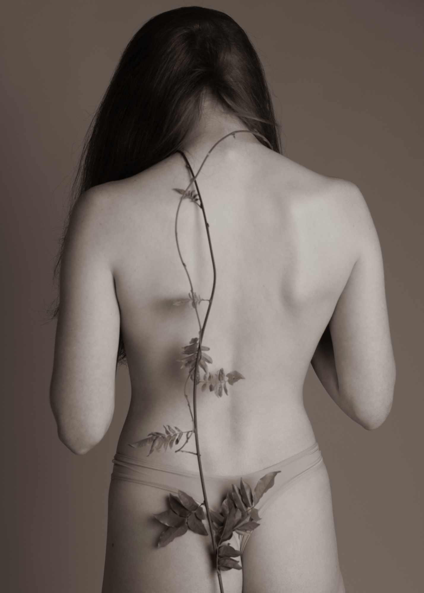 Black-and-white back view portrait of a woman with long hair and a bare back, with a climbing plant stem running down her spine.