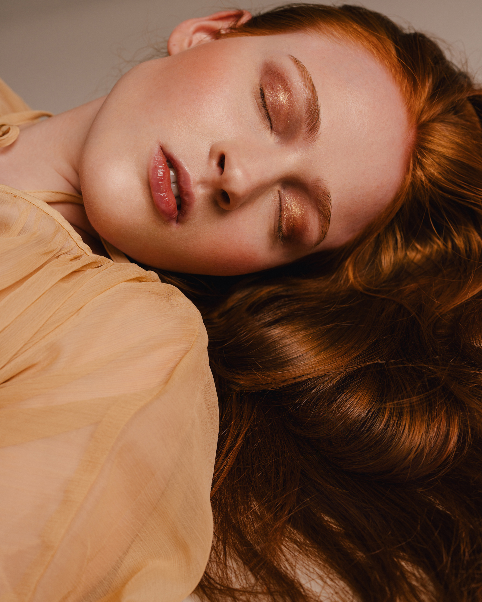 Warm-toned close-up portrait of a woman lying down with her eyes closed, surrounded by soft fabric and flowing hair.