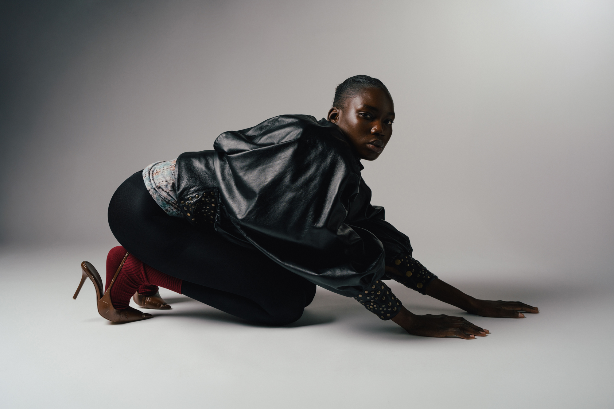 Fashion portrait of a model crouching on the studio floor, wearing a glossy black jacket and dark trousers.