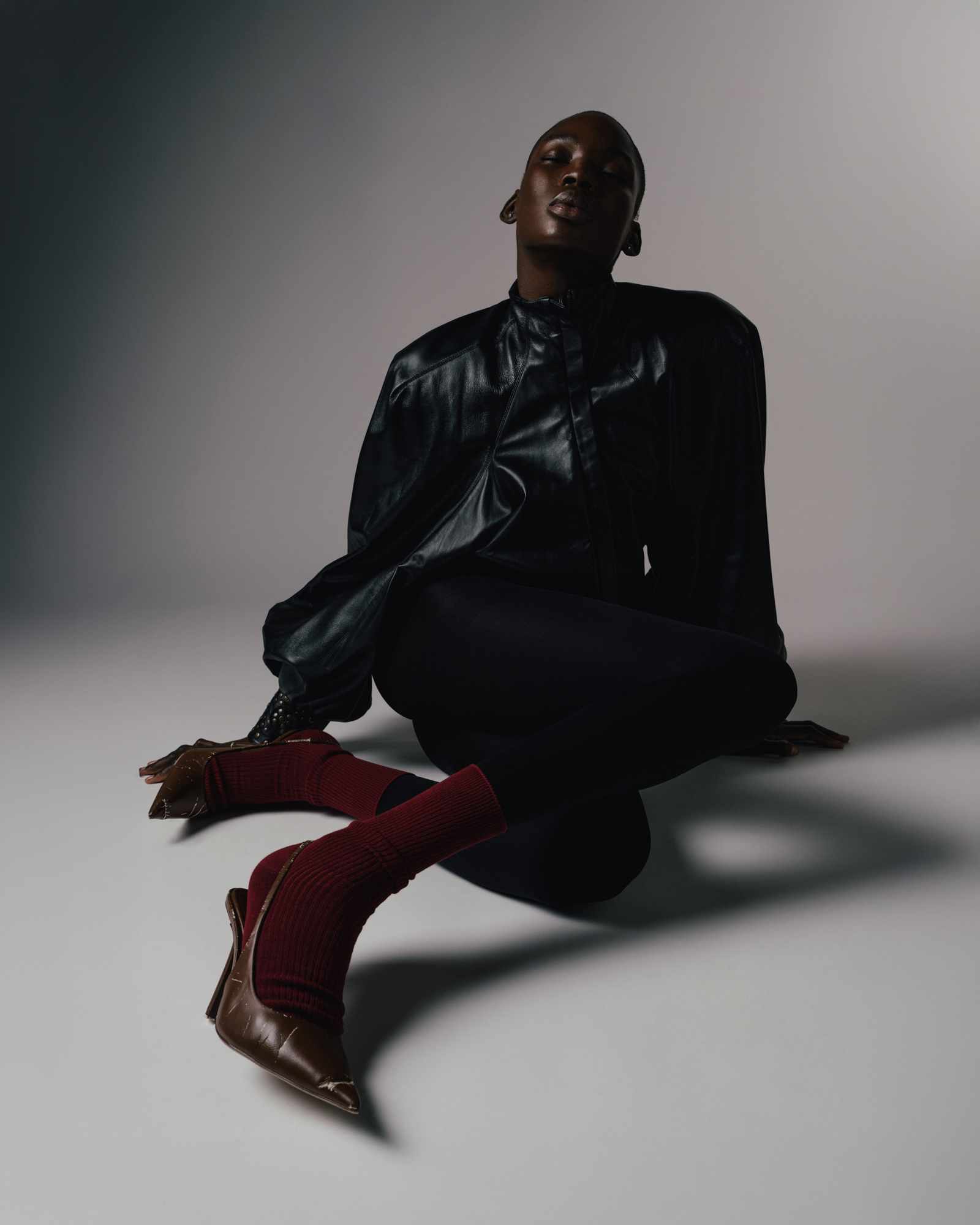 Studio fashion portrait of a seated model in black clothing and red pointed boots, leaning back against a grey backdrop.