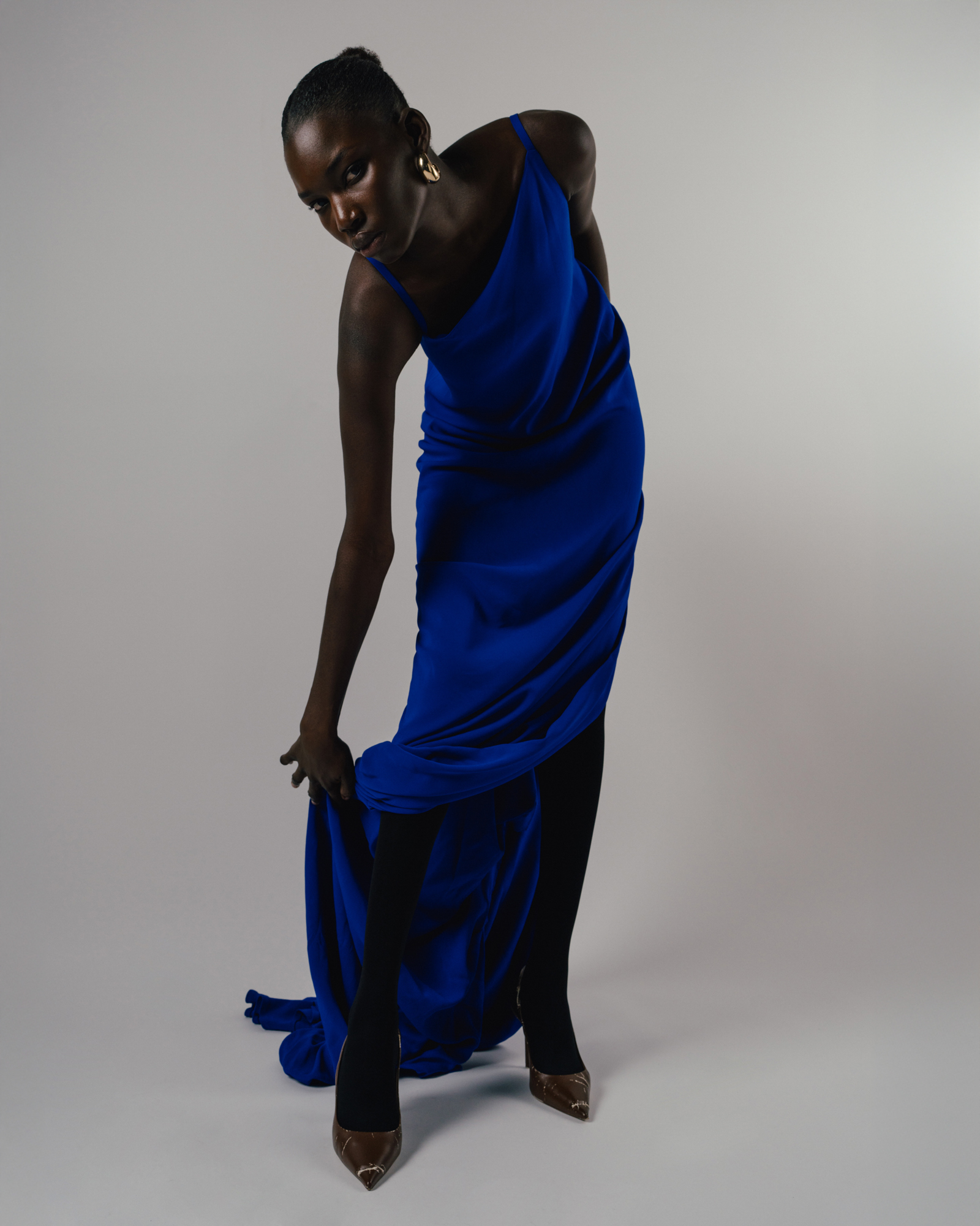 Studio fashion portrait of a model in a bright blue draped dress, bending forward and holding the hem near the floor.
