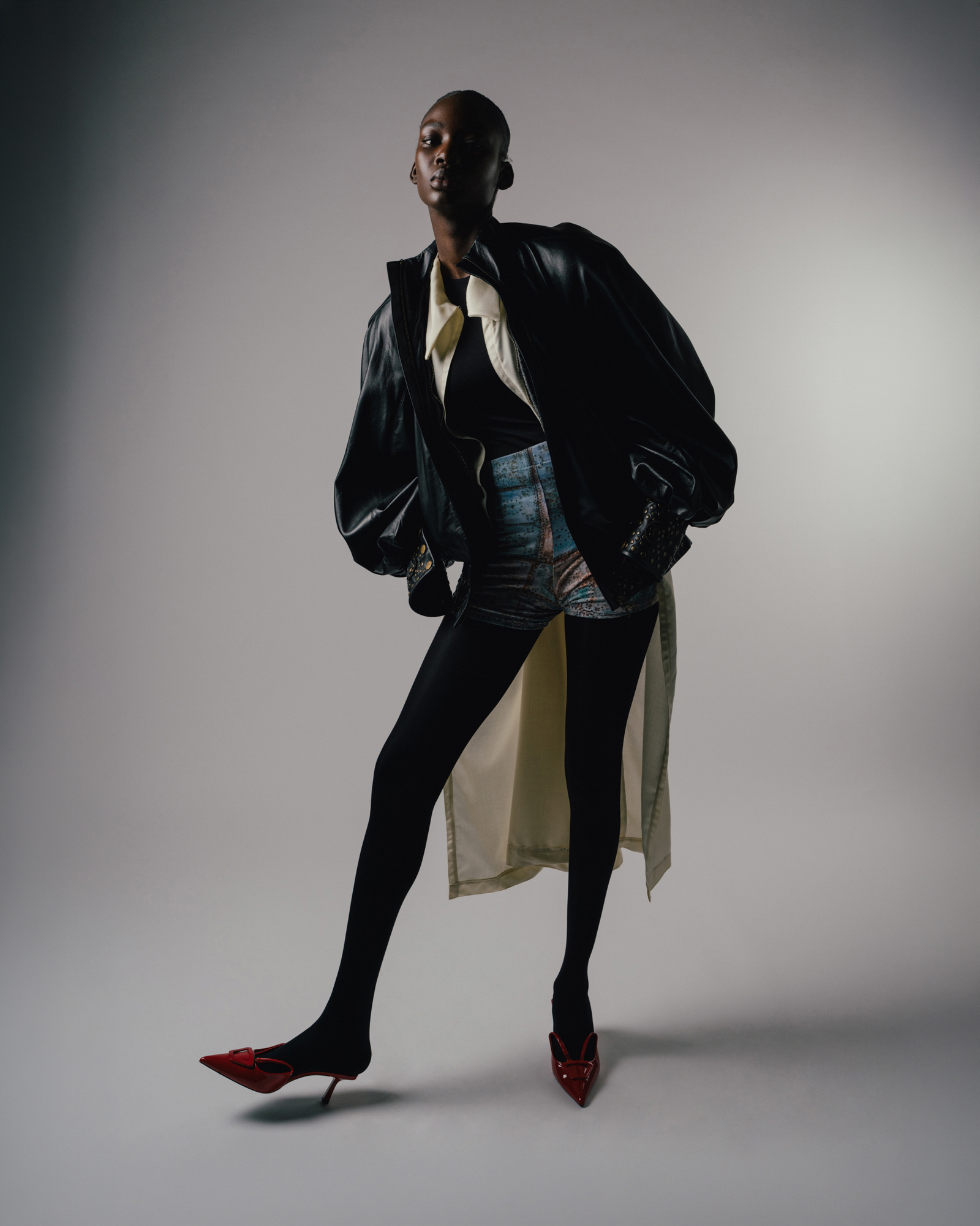 Full-body studio fashion portrait of a model wearing a black jacket, long pale skirt, and red pointed boots.