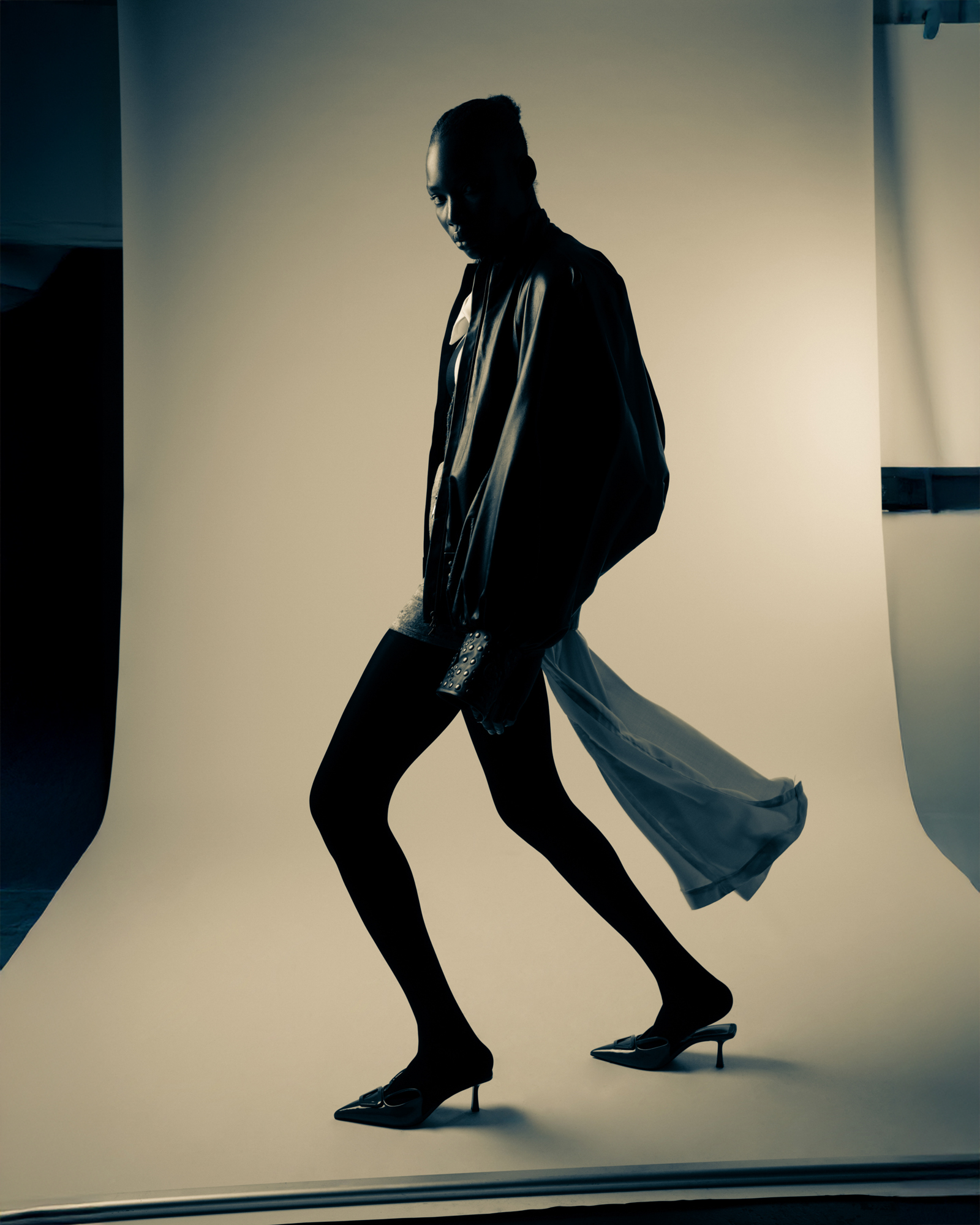 Dark studio fashion portrait of a model in layered black clothing, walking in profile with sheer fabric trailing behind.