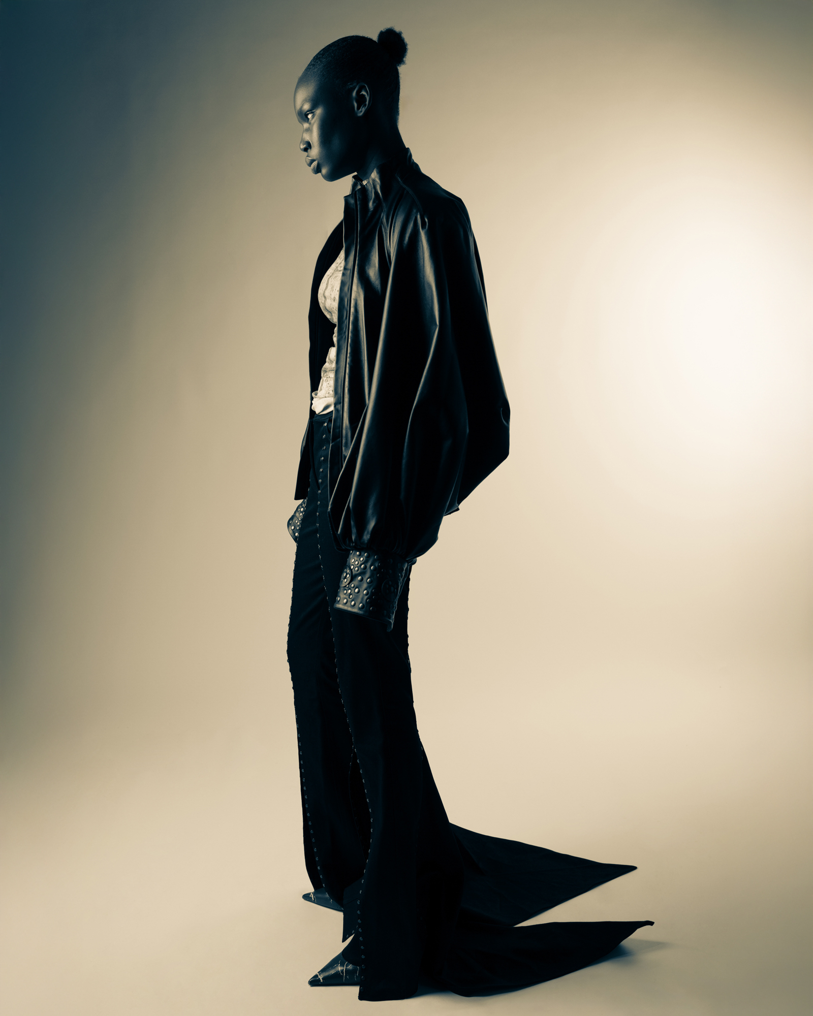 Moody studio fashion portrait of a model standing in profile, wearing a dark structured outfit with a long trailing hem.
