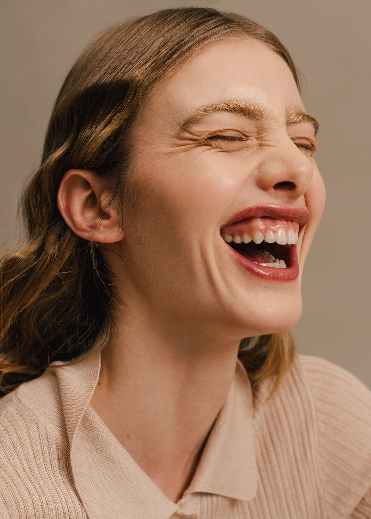 Close-up portrait of a woman laughing with her eyes closed, wearing a light knit top against a beige background.