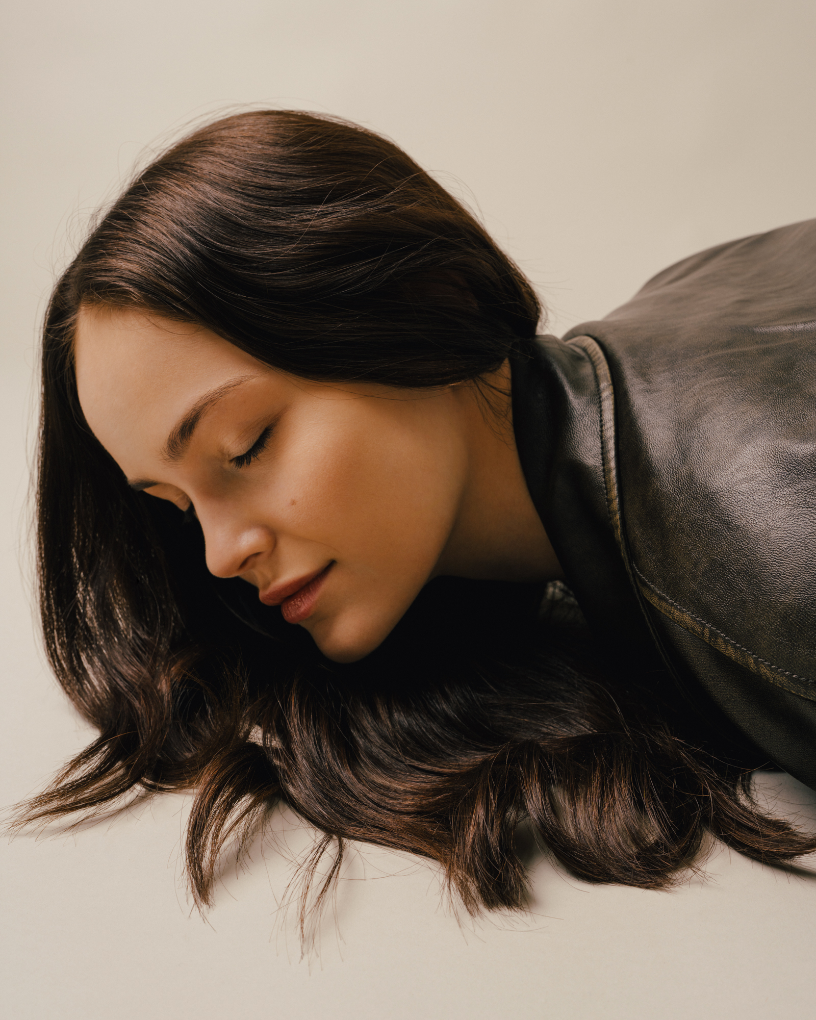 Portrait of a woman with long dark hair leaning forward in a dark textured garment against a light background.