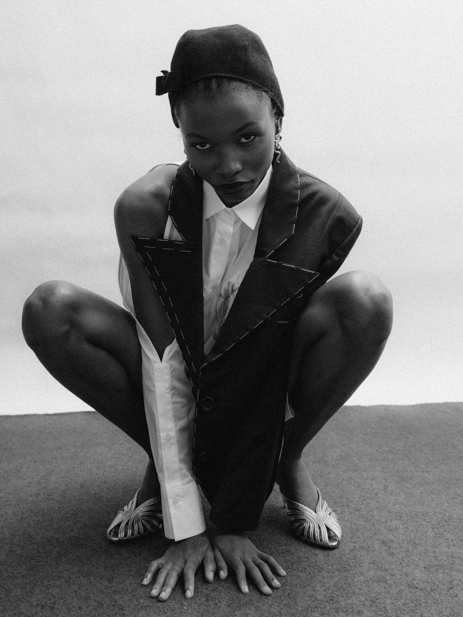 Black-and-white portrait of a crouching model in a dark vest and white shirt, posing on the floor.