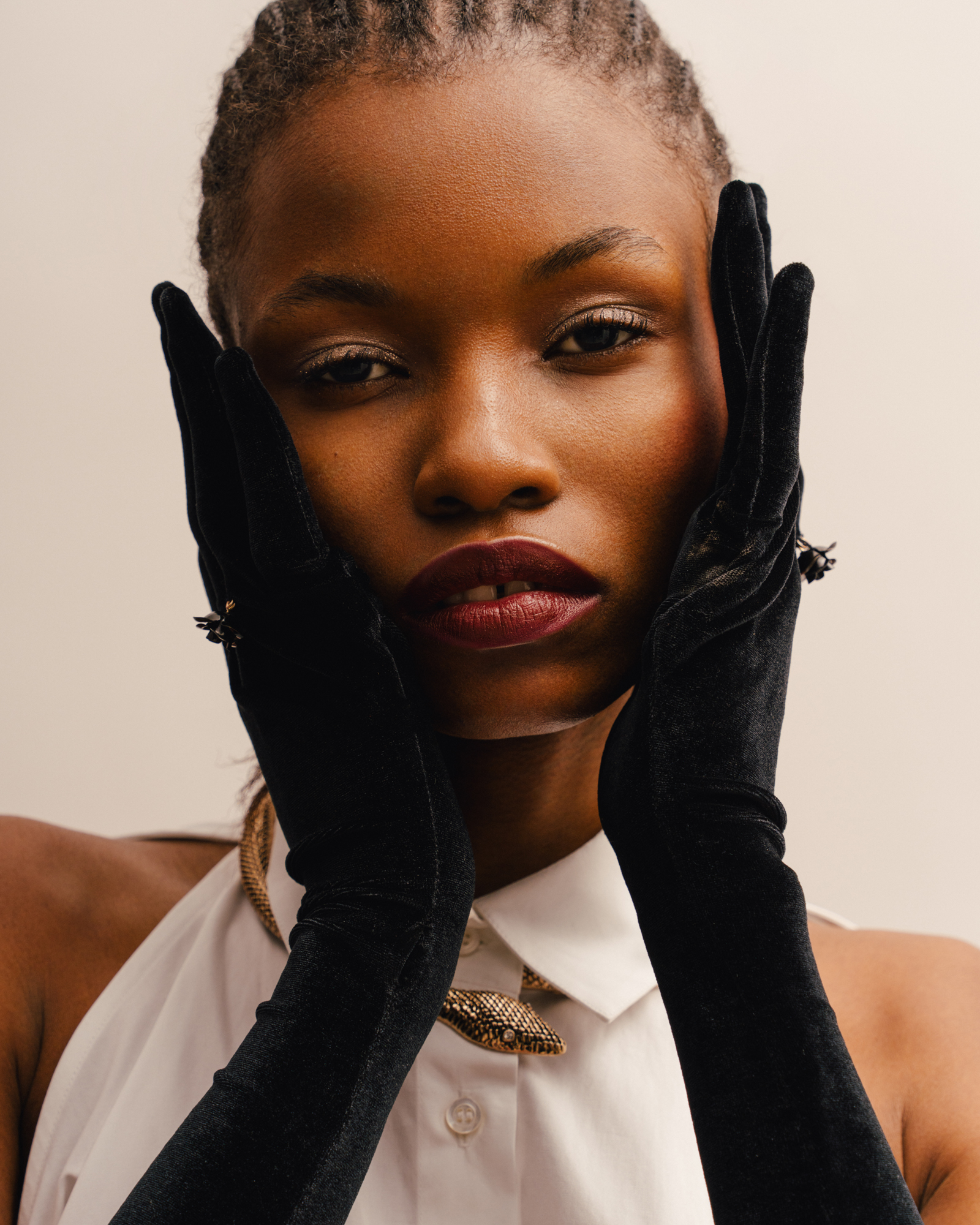 Close-up portrait of a woman wearing long black gloves, framing her face with her hands against a light background.