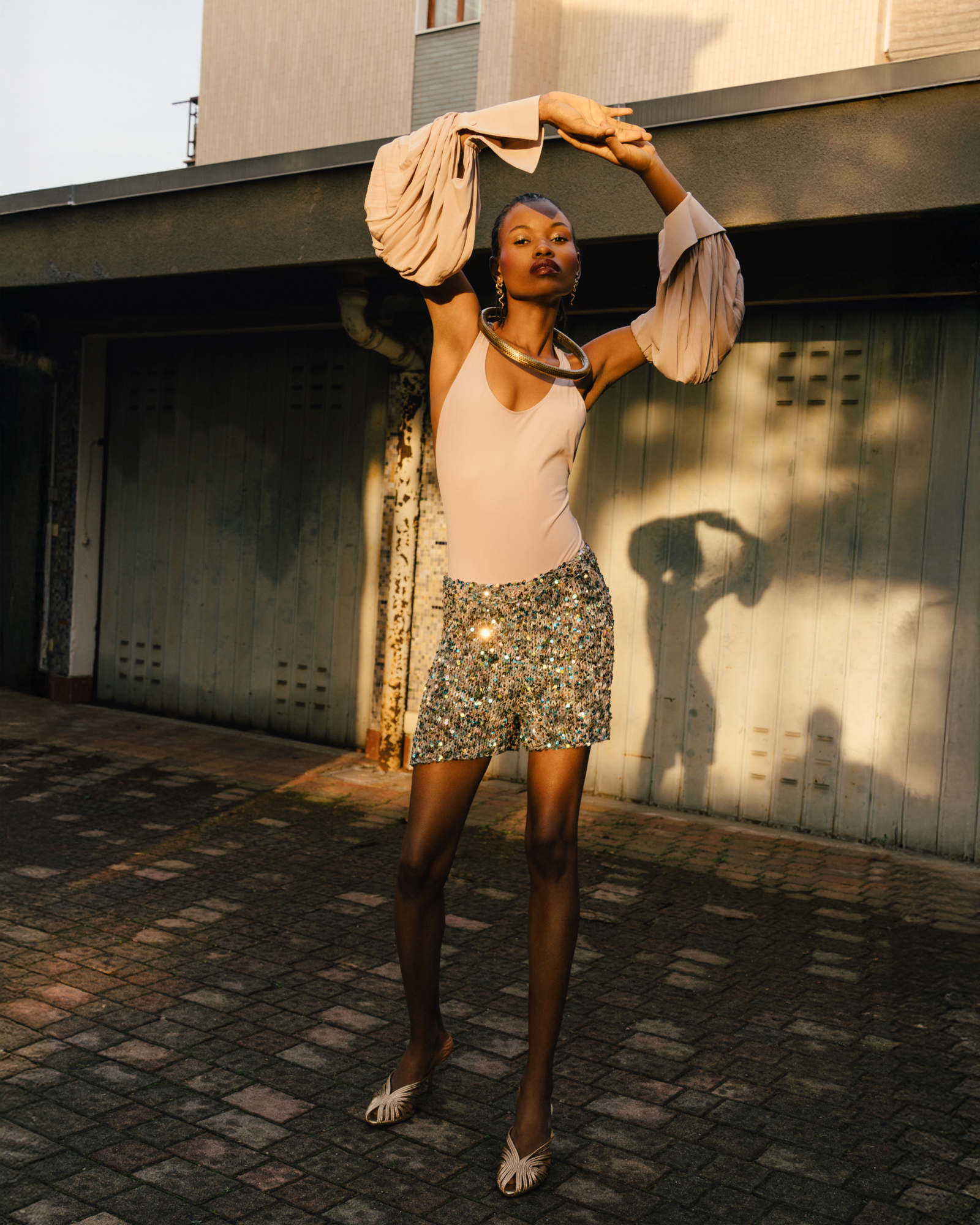 Fashion portrait of a model standing outdoors in a pale top, patterned skirt, wide-brim hat, and high heels.