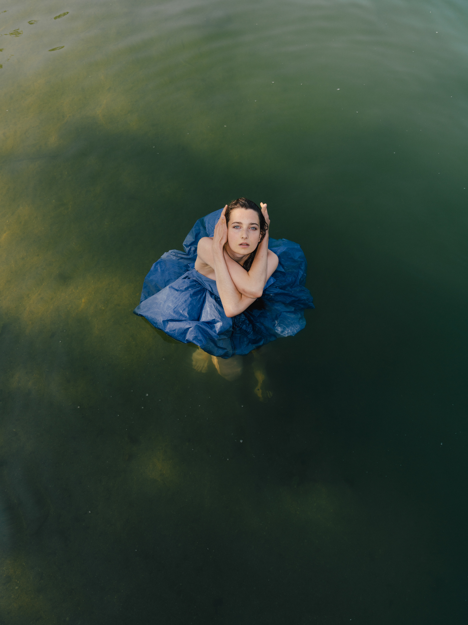 Portrait of a woman floating in dark green water, wearing a voluminous blue garment.