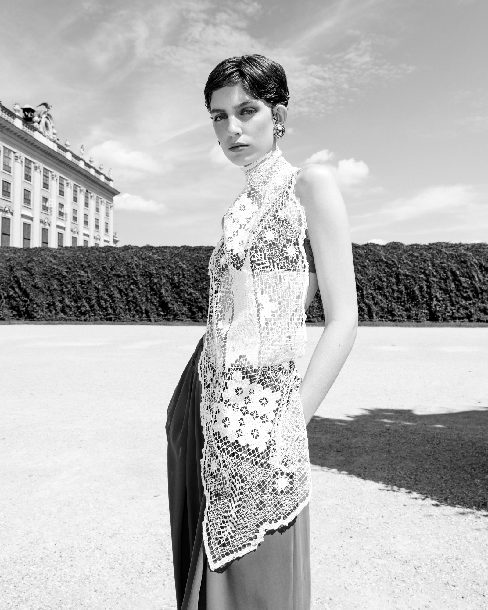Black-and-white outdoor fashion portrait of a woman in a patterned sleeveless dress, standing in front of a historic building and garden.