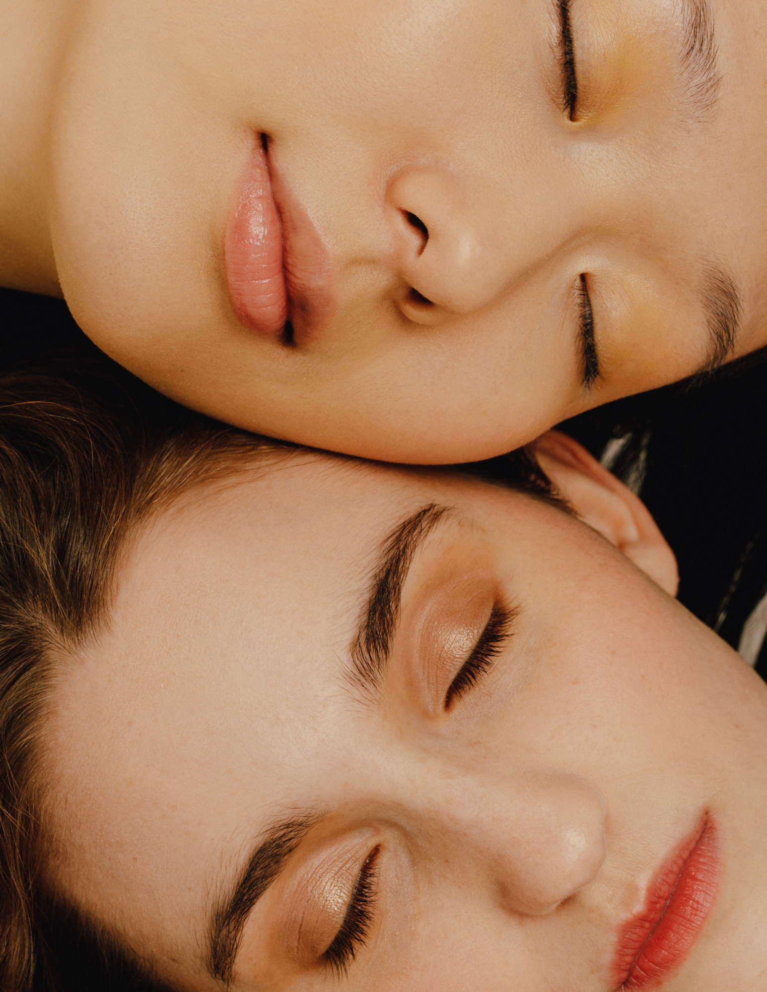 Close-up portrait of two women with their eyes closed, their faces resting gently against each other in soft, natural light.