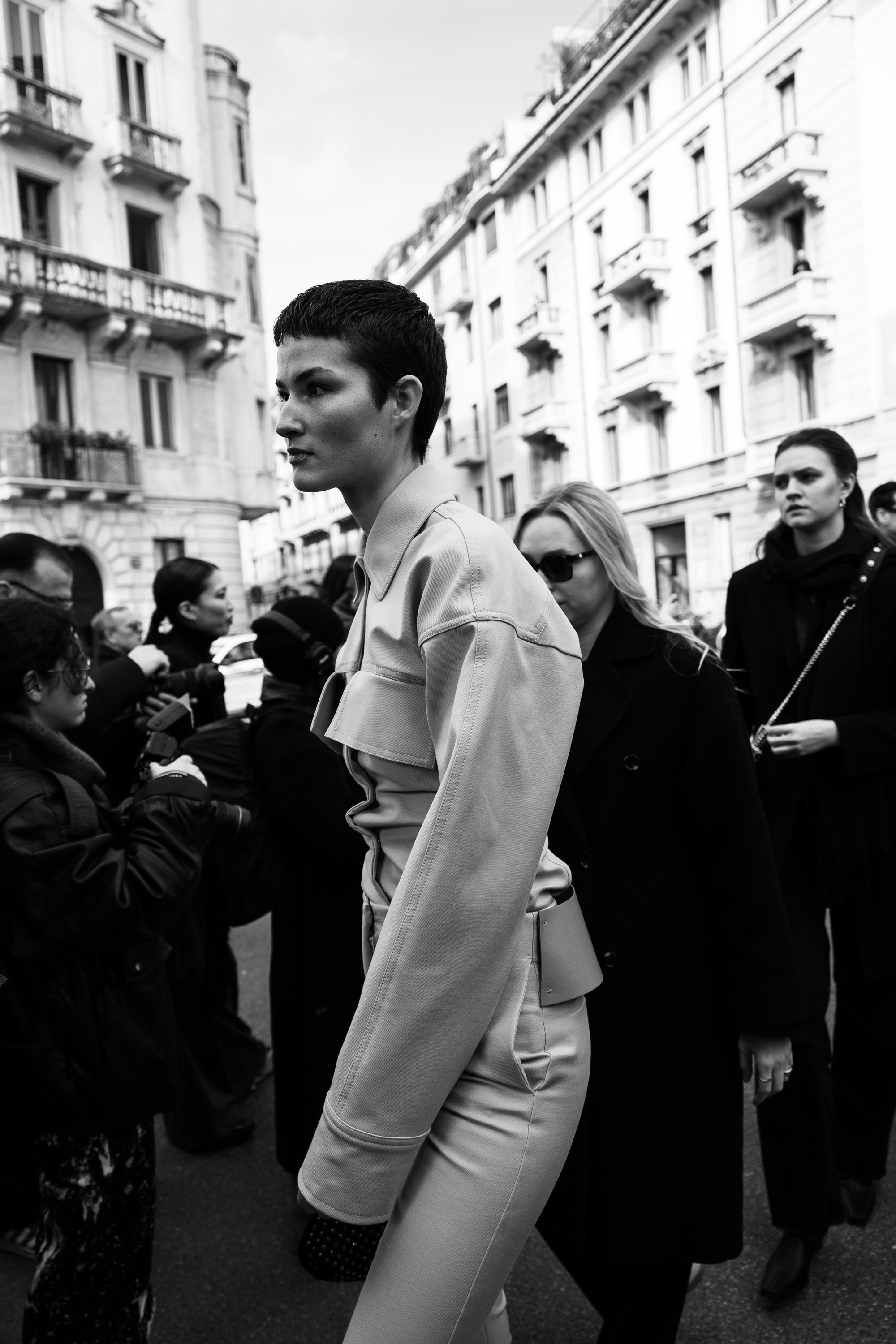 A black-and-white street photo of a person with short hair wearing a structured light-colored outfit, walking through a crowd outside historic city buildings.