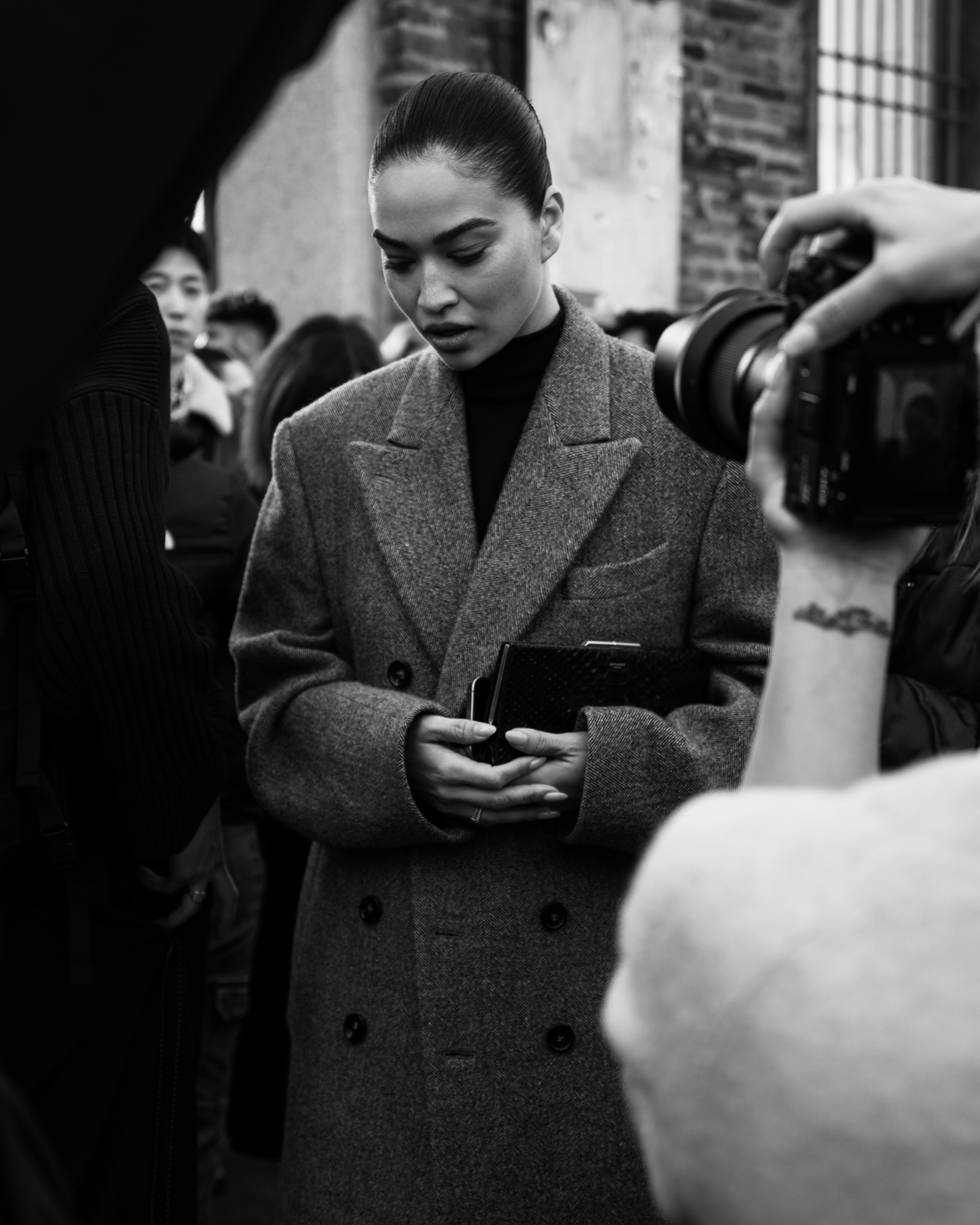 A black-and-white candid photo of a person in an oversized double-breasted coat holding a clutch, surrounded by photographers and a crowd outside a brick building.