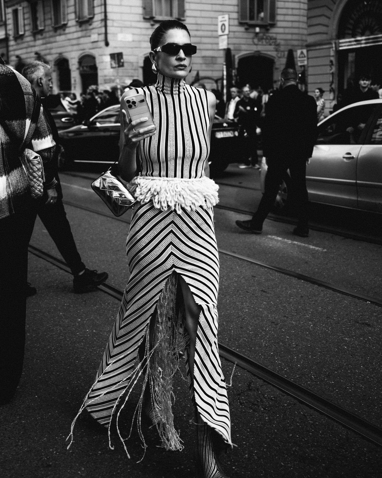 A black-and-white street style photo of a person wearing sunglasses and a striped sleeveless dress with a textured white waist detail, standing on a city street while holding a smartphone.