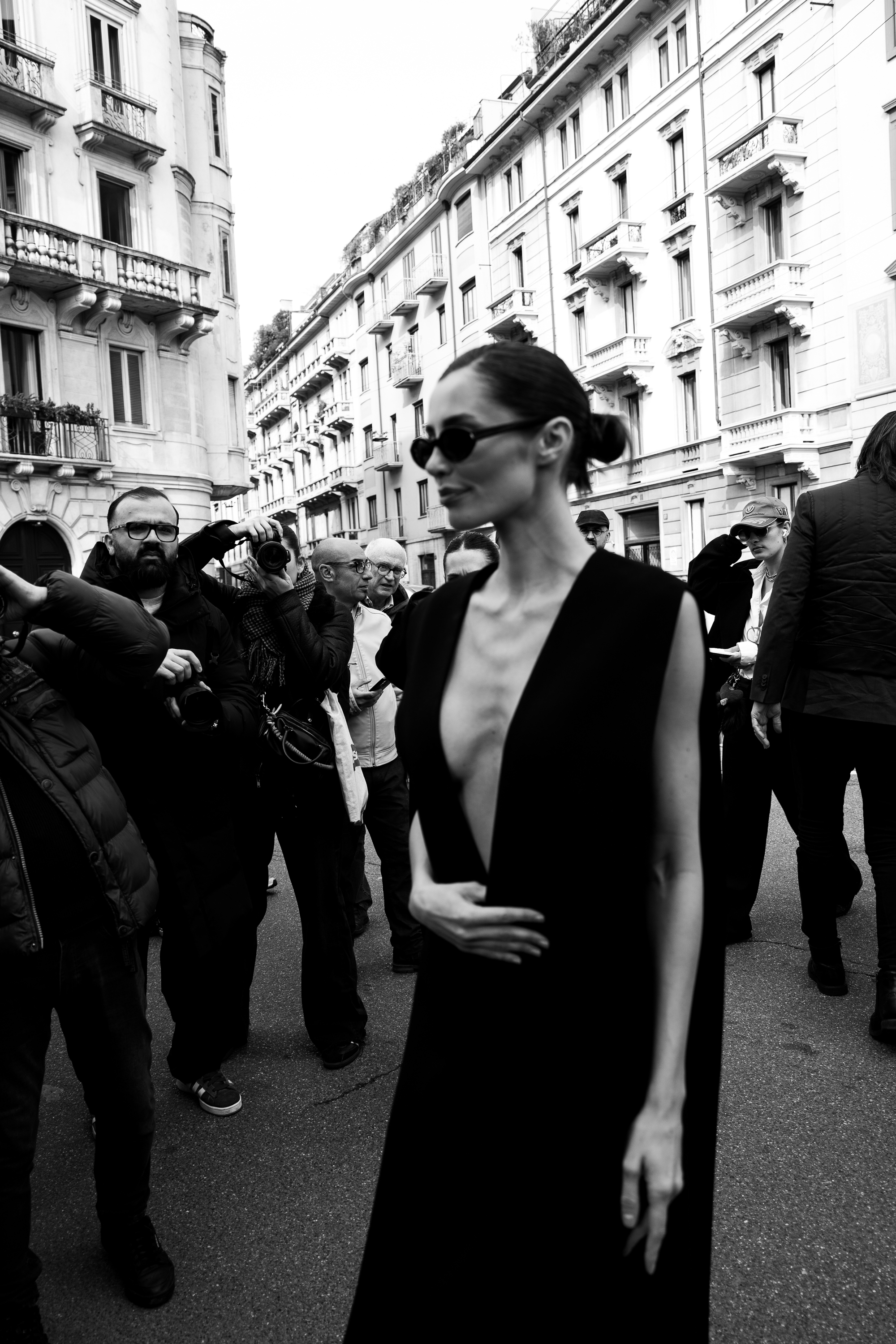 A black-and-white street photo of a person in sunglasses and a deep V-neck sleeveless dress walking past photographers on a city street lined with historic buildings.
