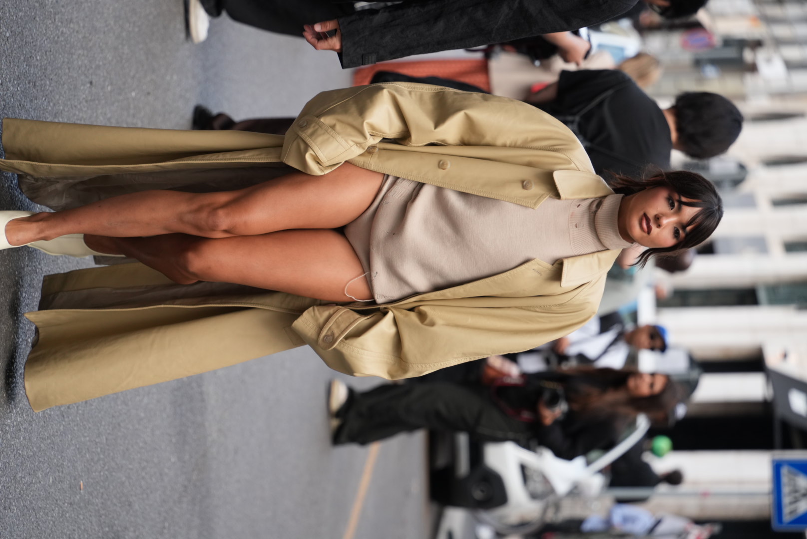Woman wearing an oversized beige trench coat over a taupe turtleneck bodysuit, walking on a city street with her hands in the coat pockets.