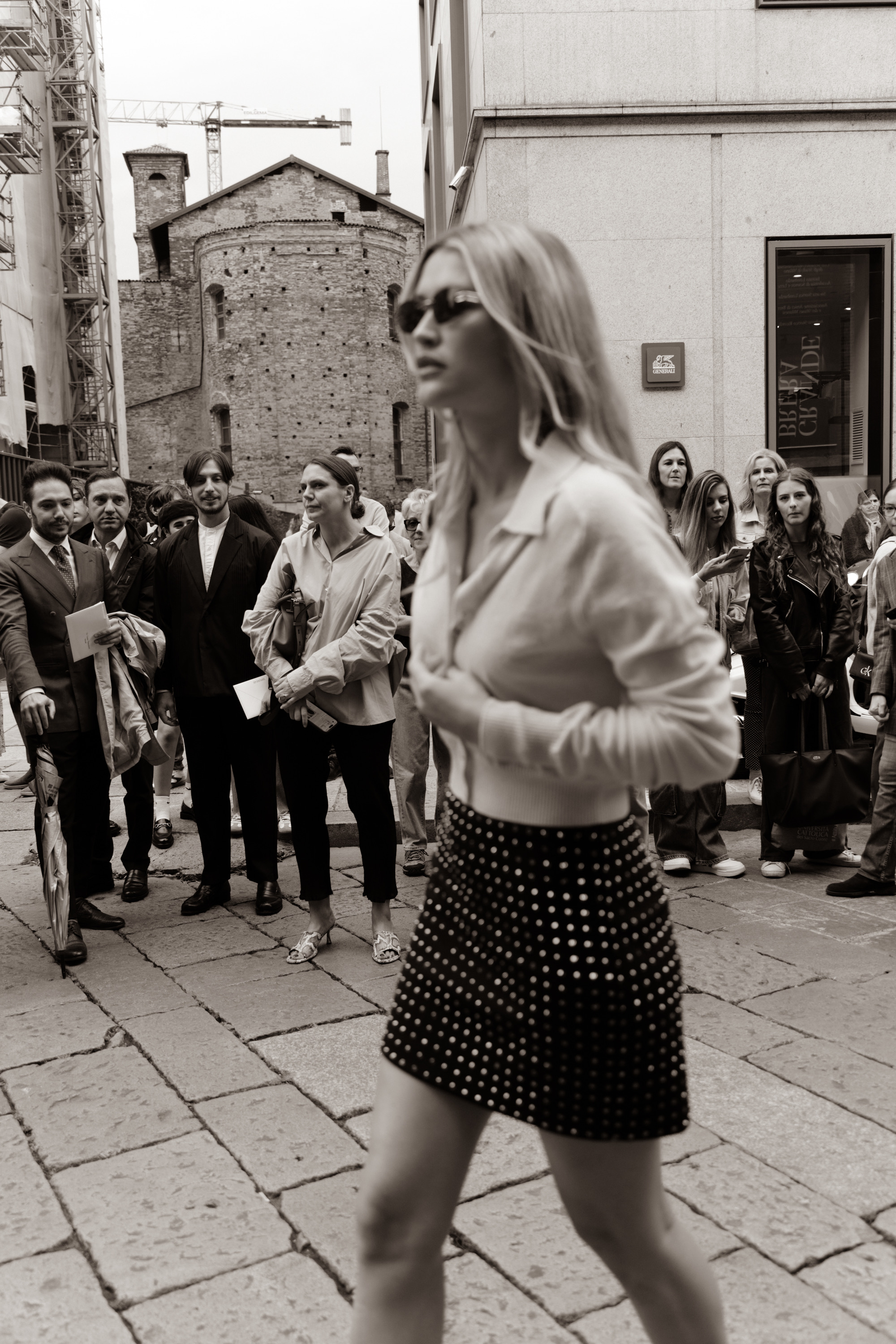 A black-and-white street style photo of a person wearing sunglasses, a white blouse, and a black polka-dot mini skirt, walking past a crowd in an urban courtyard.
