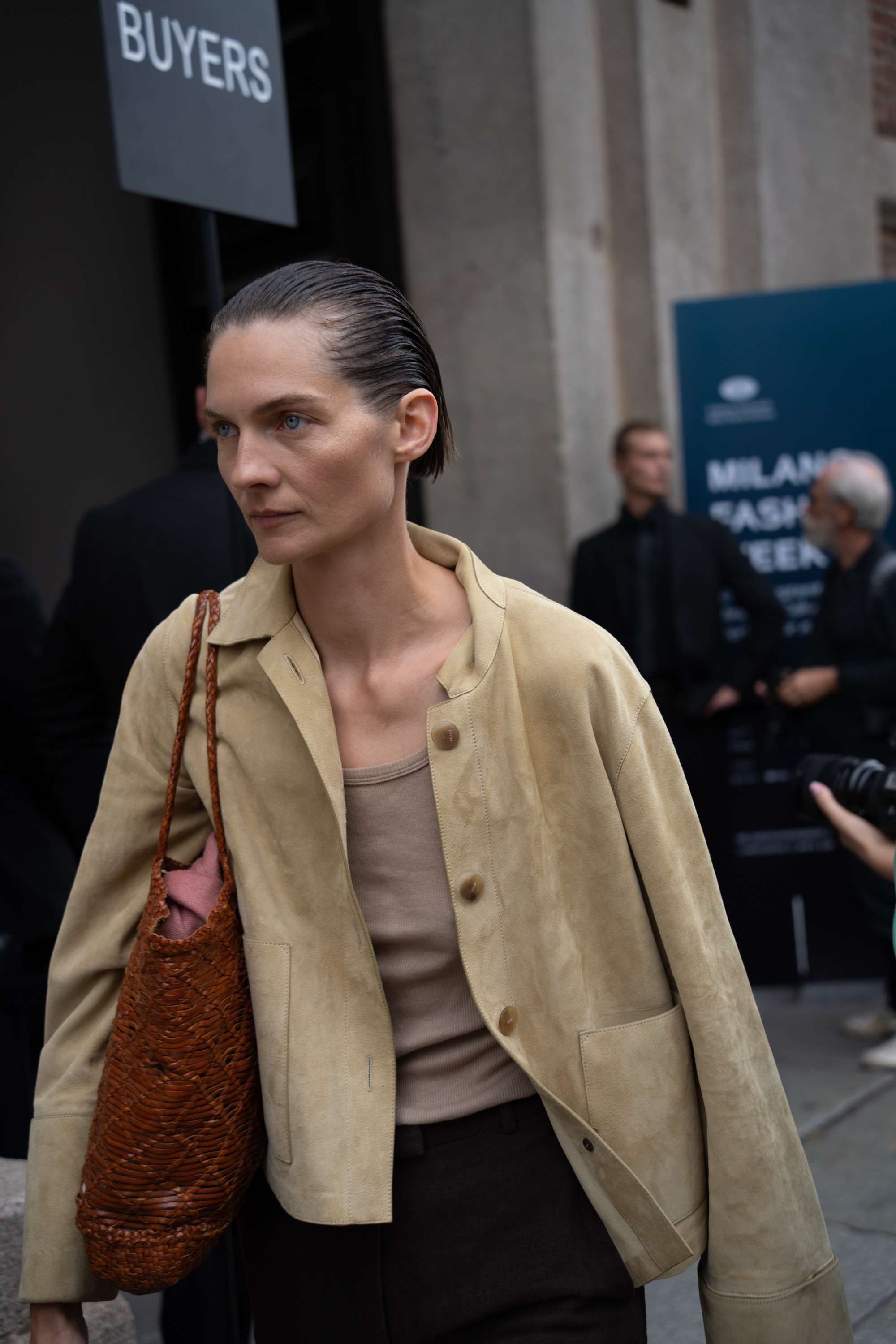 A street style photo of a person with slicked-back hair wearing sunglasses, a cropped beige jacket, and a woven brown shoulder bag outside a venue entrance.