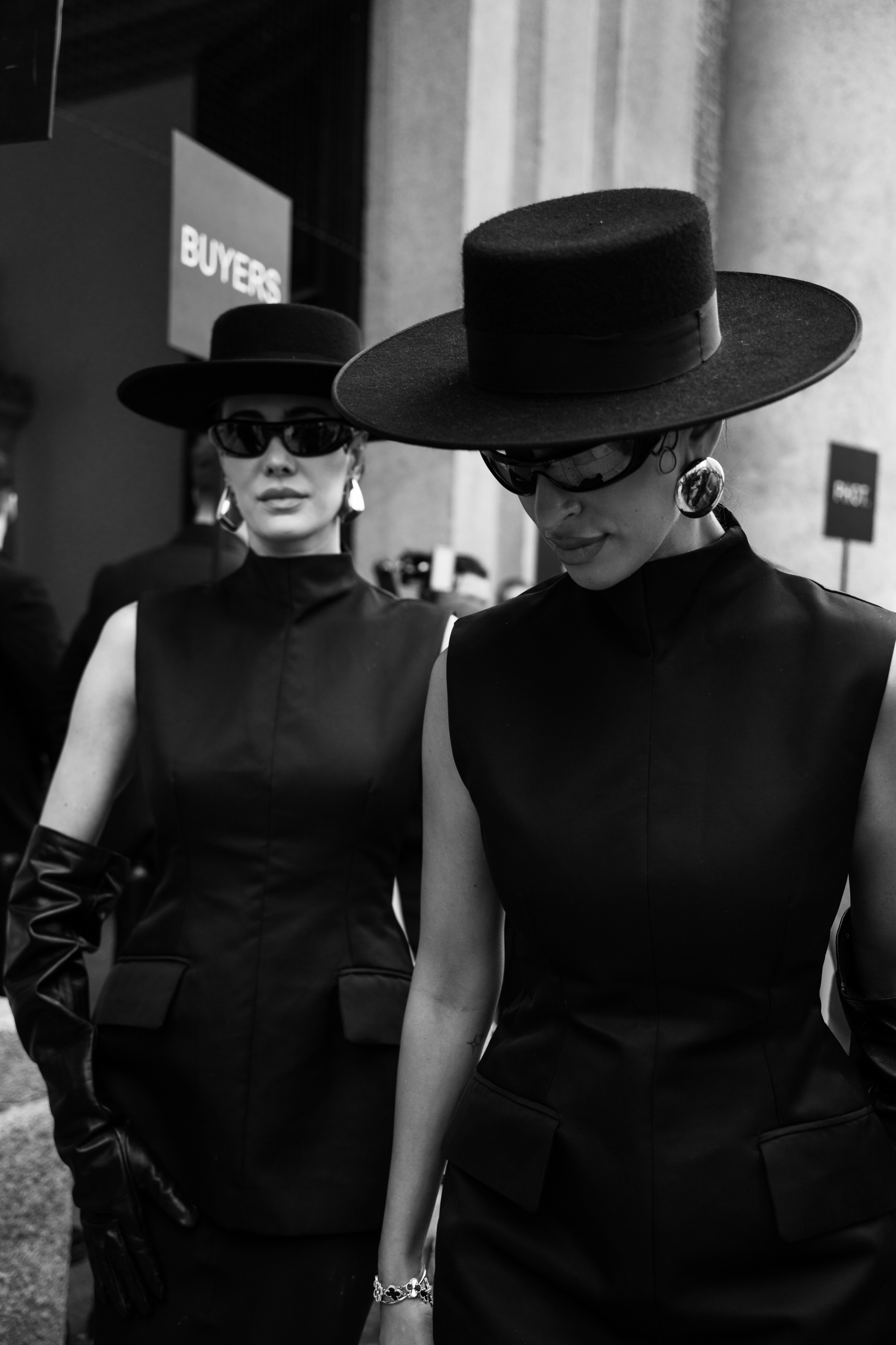 A black-and-white street style photo of two people wearing wide-brimmed black hats, sunglasses, and sleeveless black structured outfits outside a venue entrance.