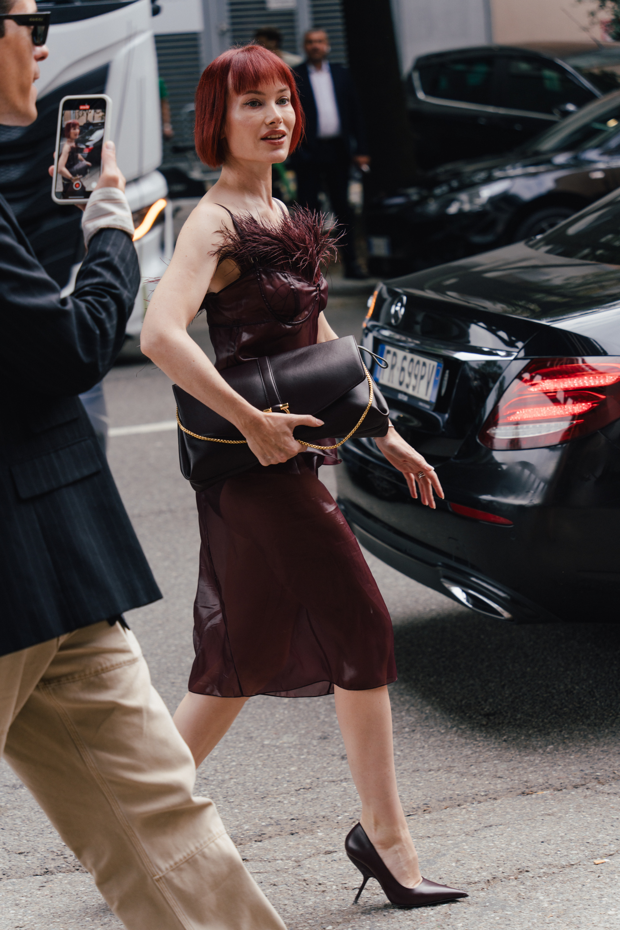 A street style photo of a person with a red bob haircut wearing a burgundy dress with feather detailing, walking beside a black car while holding a dark clutch.