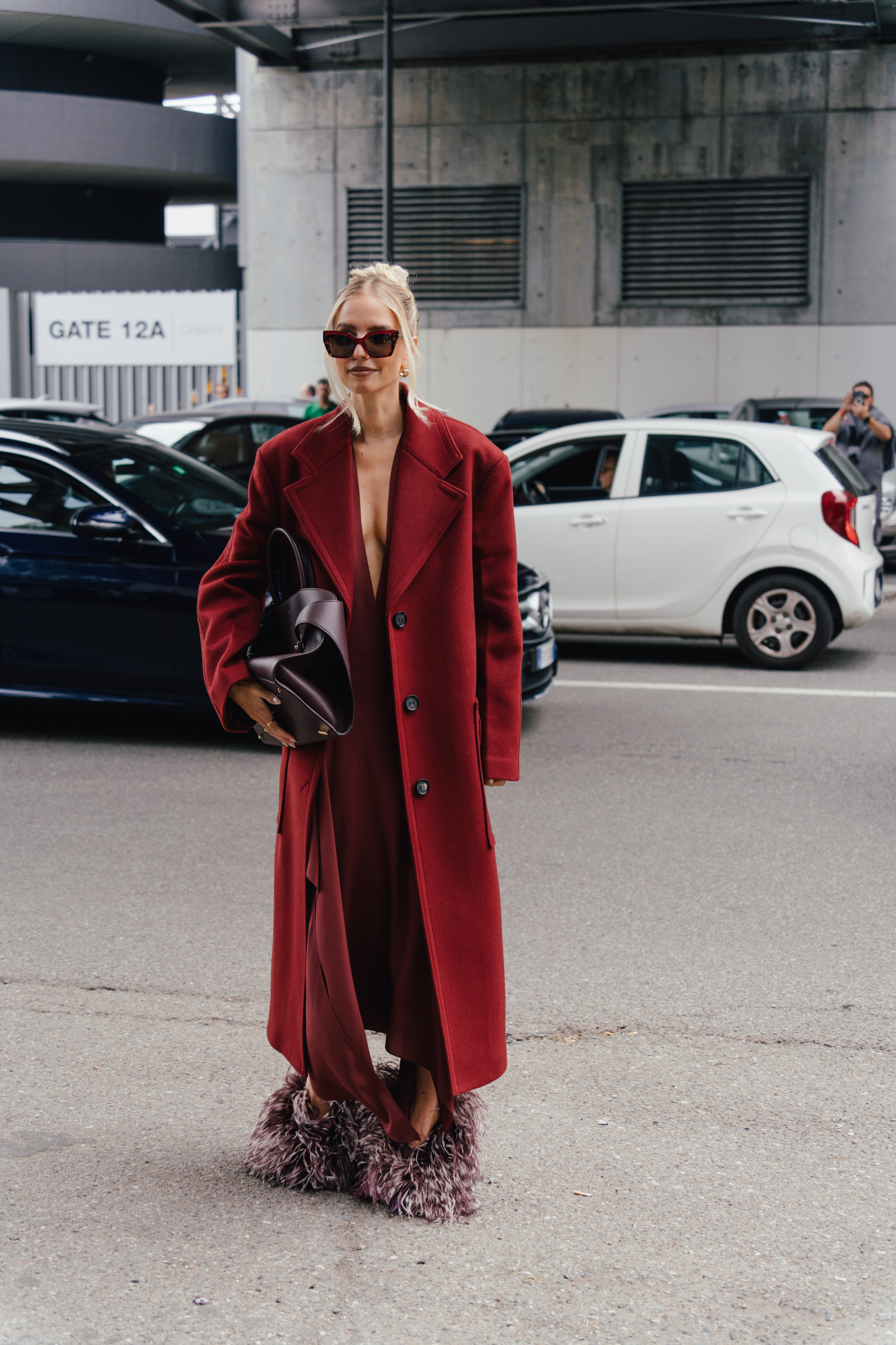 A street style photo of a person wearing sunglasses, a long red coat, and fuzzy purple shoes, standing near a gate sign while holding a large dark clutch.