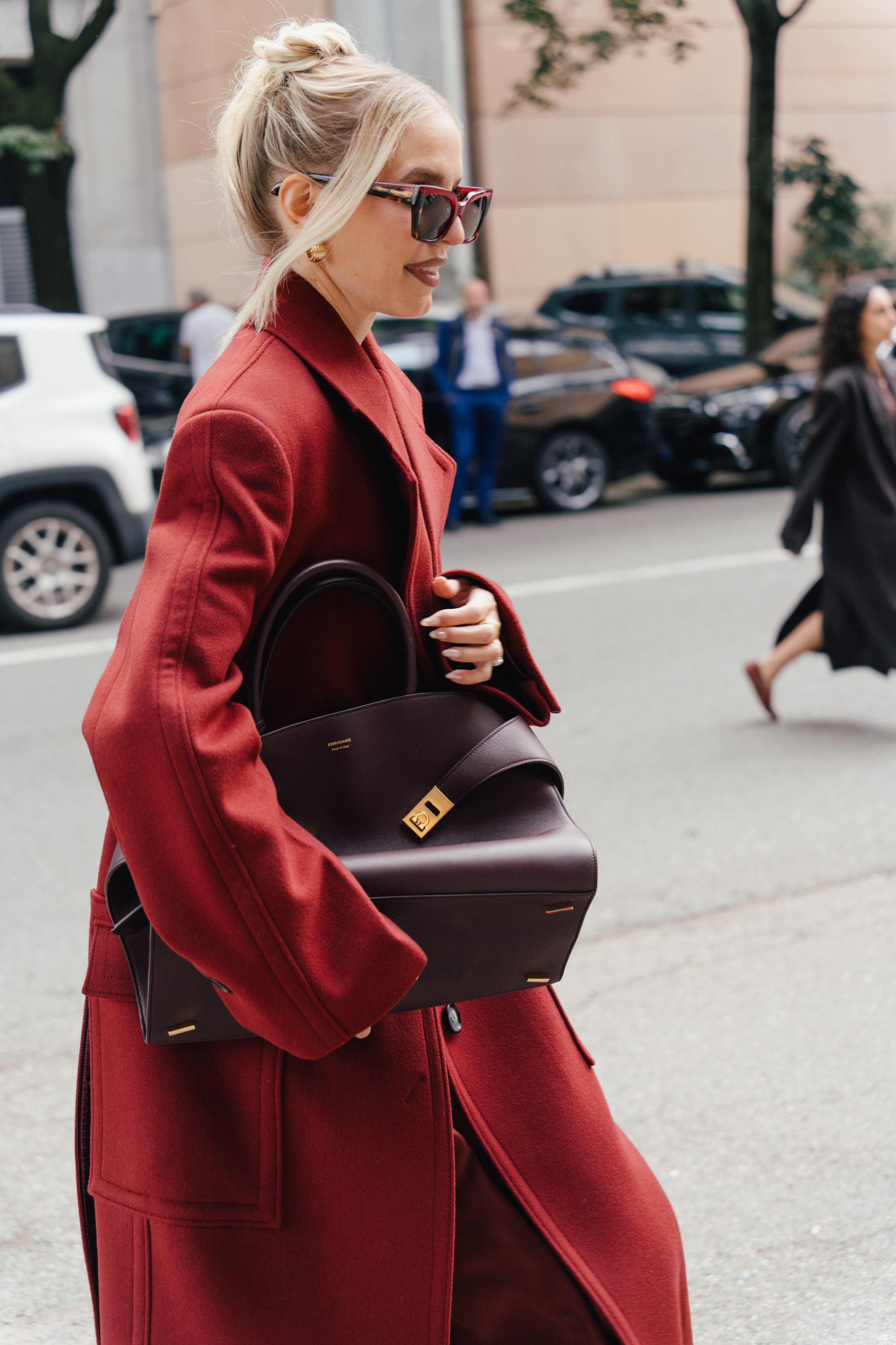 A close-up street style photo of a person in red sunglasses and a long red coat, walking while carrying a large dark structured handbag.