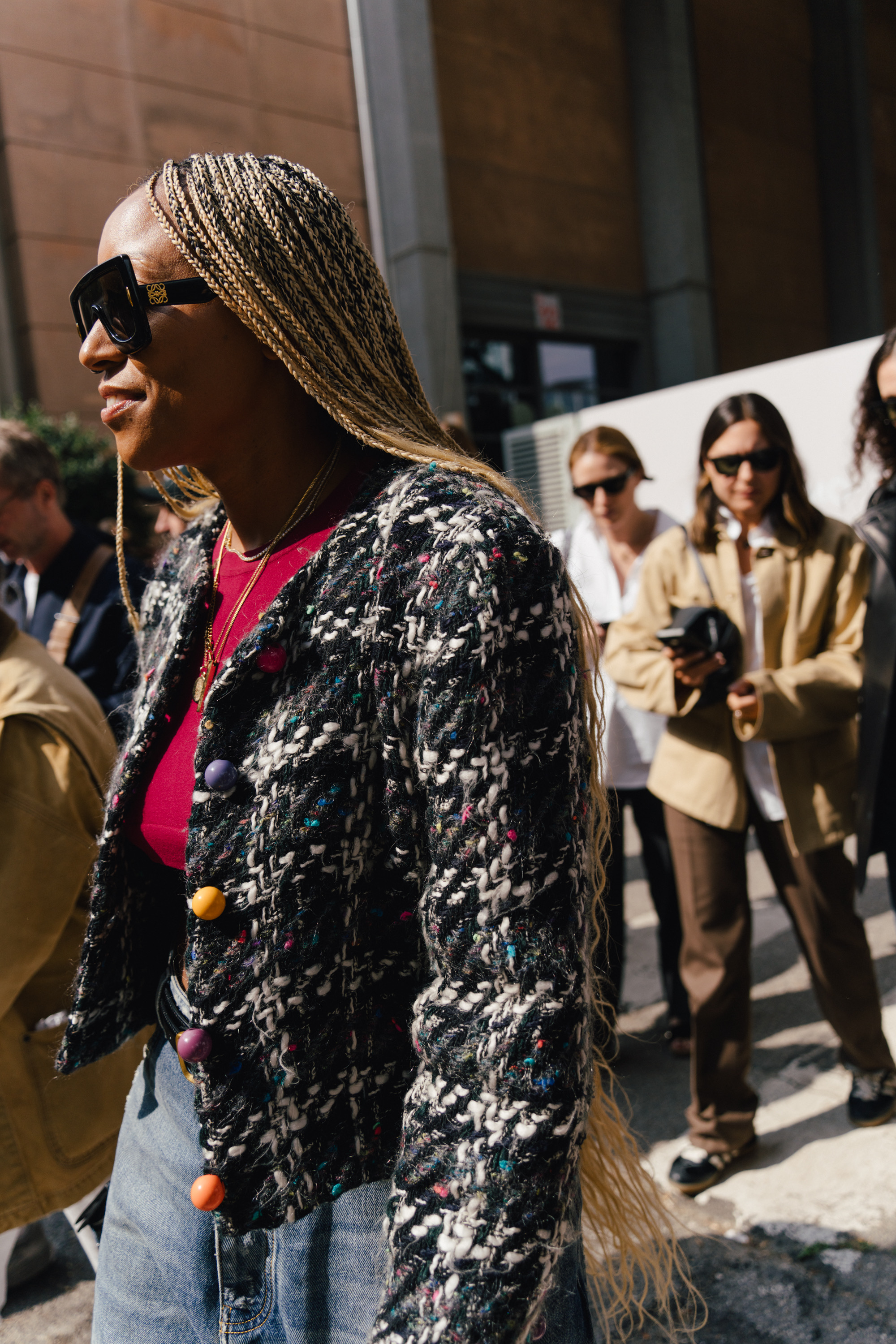 A street style photo of a person wearing sunglasses, long blonde braids, a textured black-and-white jacket with colorful buttons, and a bright pink top, standing in a crowd on a sunny city street.